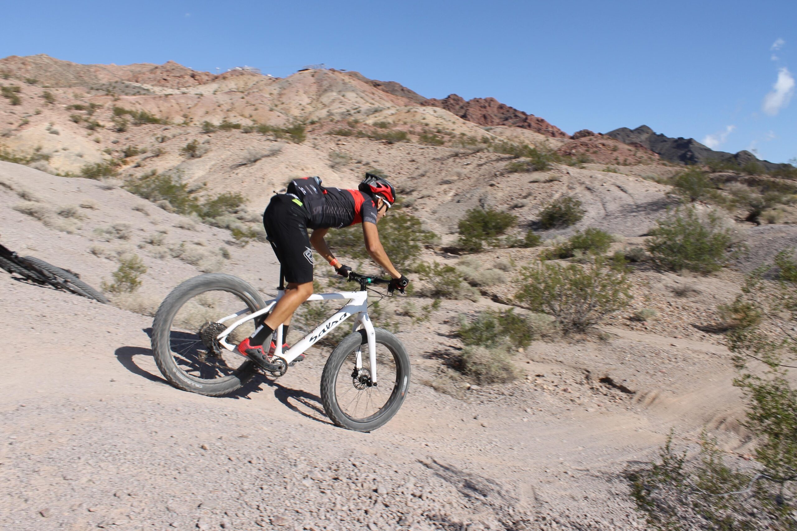 A person riding a mountain bike on a rocky, desert trail, with a backdrop of rugged hills and clear blue skies. The cyclist is in a forward-leaning position, wearing a red and black cycling outfit and a helmet, navigating a downhill path surrounded by sparse vegetation and gravel. Bootleg Canyon mountain bike trail.