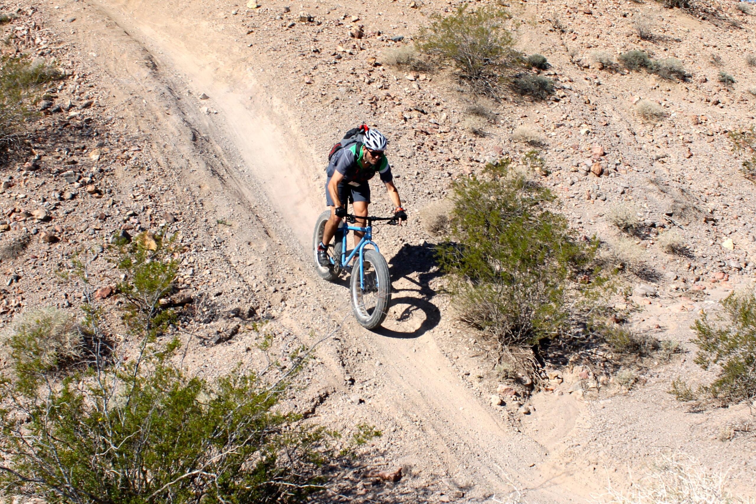 A cyclist riding a blue fat bike on a dirt trail in a rocky desert landscape, with scattered bushes and dry terrain. Dust is being kicked up from the bike tires as the cyclist navigates the uneven path. Bootleg Canyon mountain bike trail.