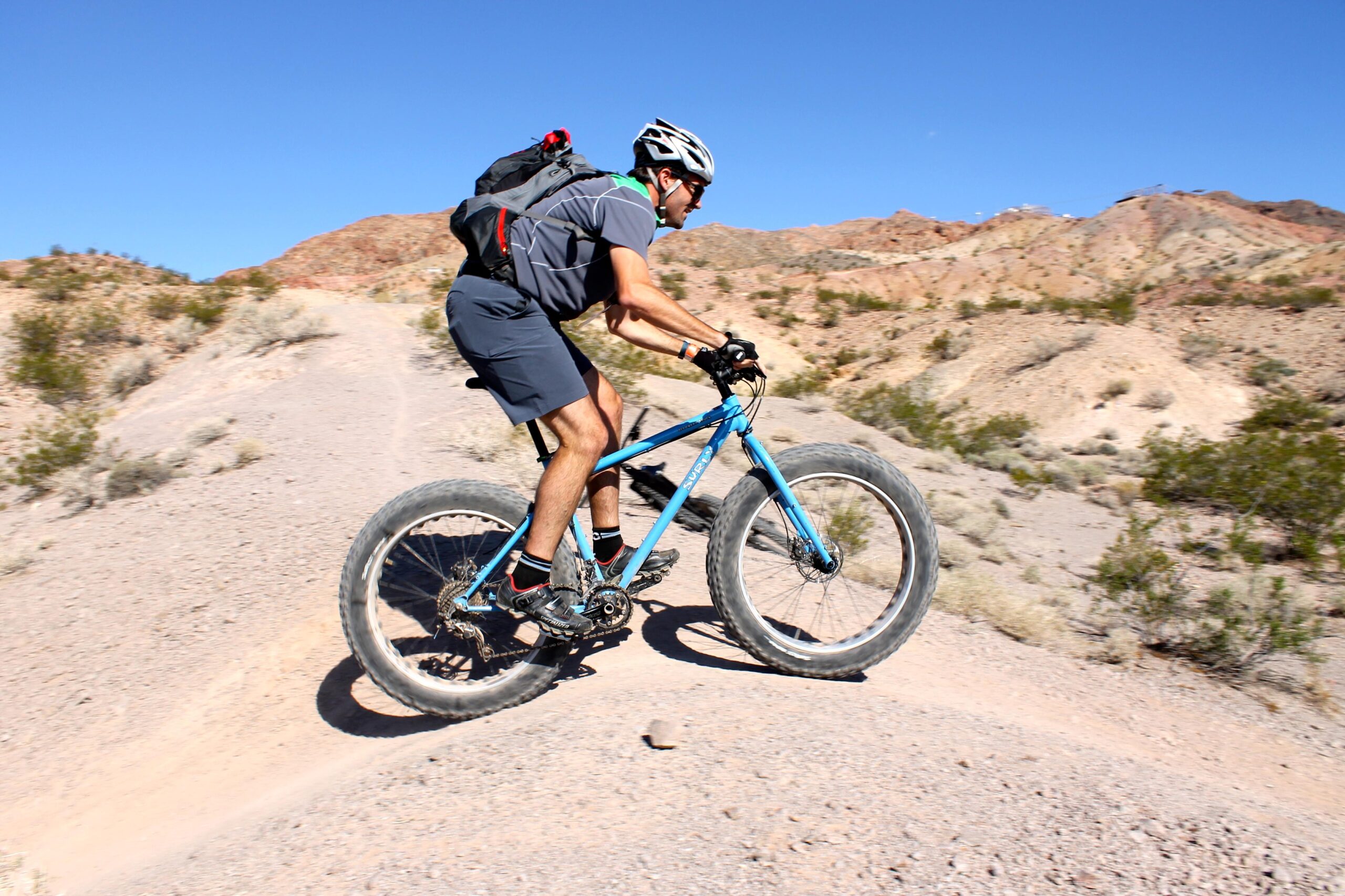 A mountain biker riding on a dirt trail, navigating up an incline in a rugged, desert landscape. The bike is blue and features wide tires, and the rider is wearing a helmet and sports gear, with a backpack visible. The background showcases rocky hills under a clear blue sky. Bootleg Canyon mountain bike trail.