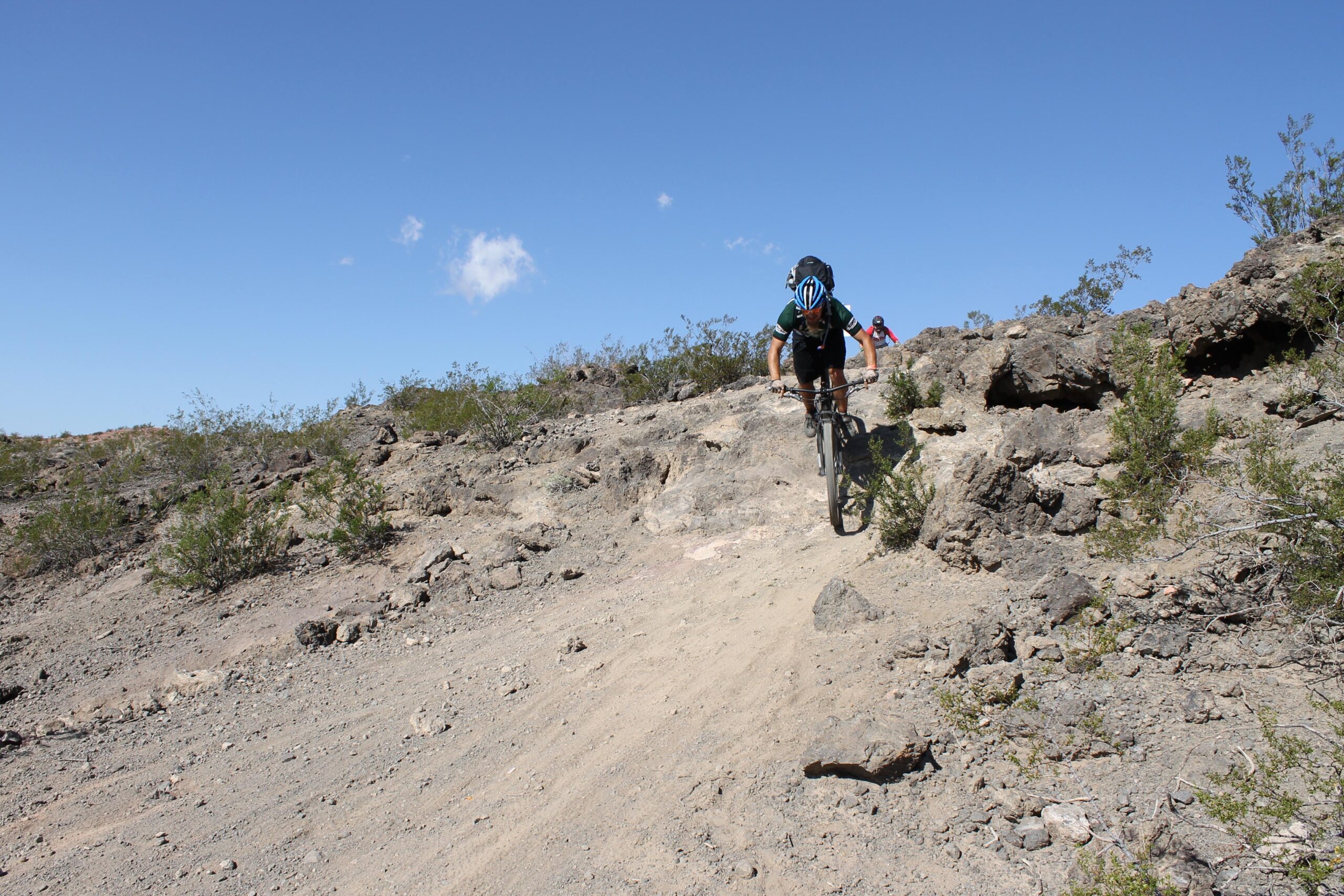 A person riding a mountain bike on a rocky trail in a desert landscape under a clear blue sky, with sparse vegetation in the foreground and a second rider visible in the background. Bootleg Canyon mountain bike trail.