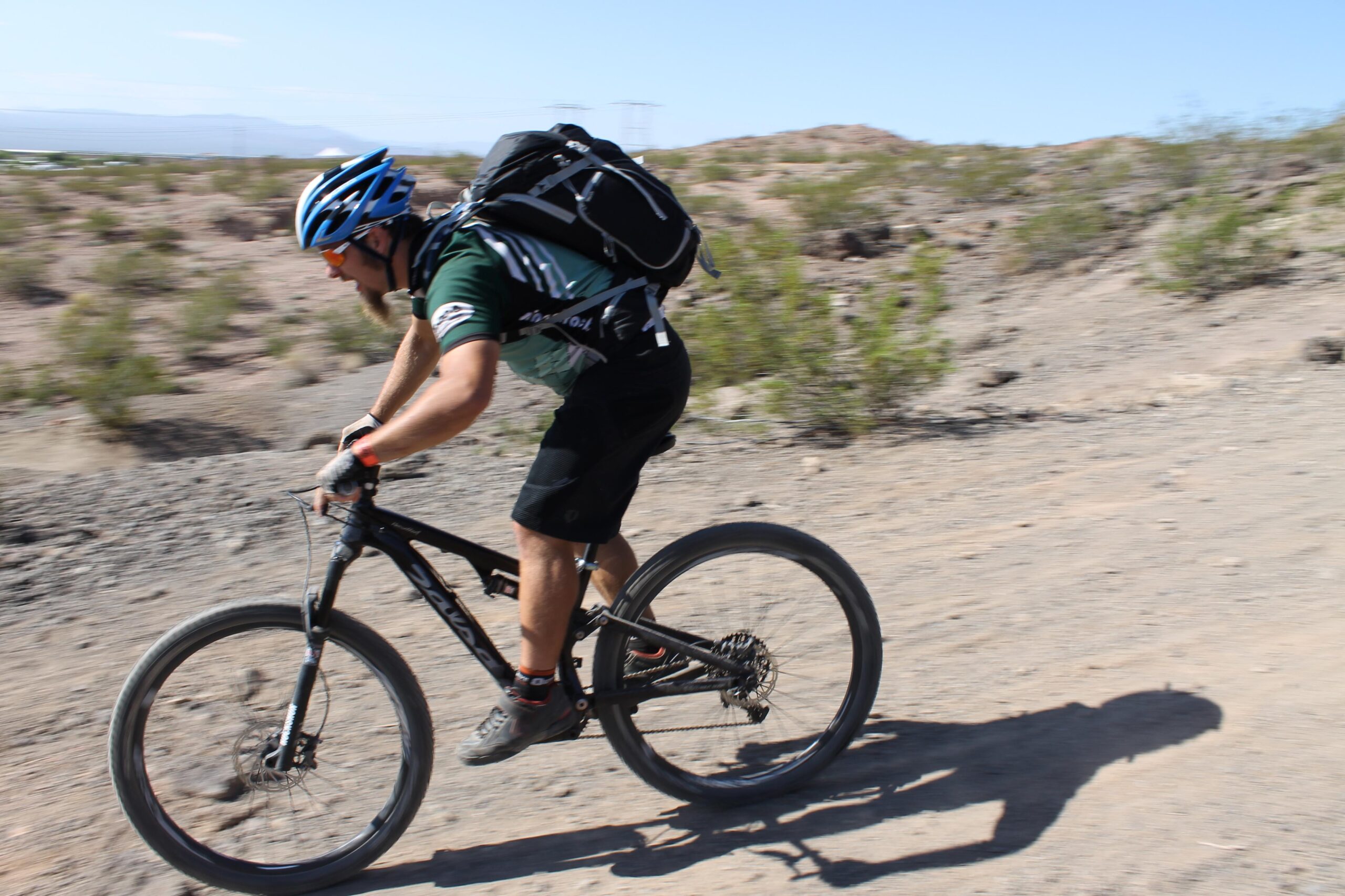 A mountain biker riding on a dirt trail in a desert landscape. The cyclist is wearing a blue helmet and a backpack, leaning forward for speed, with blurred motion effect indicating brisk movement. Sparse vegetation and rocky terrain are visible in the background under a clear blue sky. Bootleg Canyon mountain bike trail.