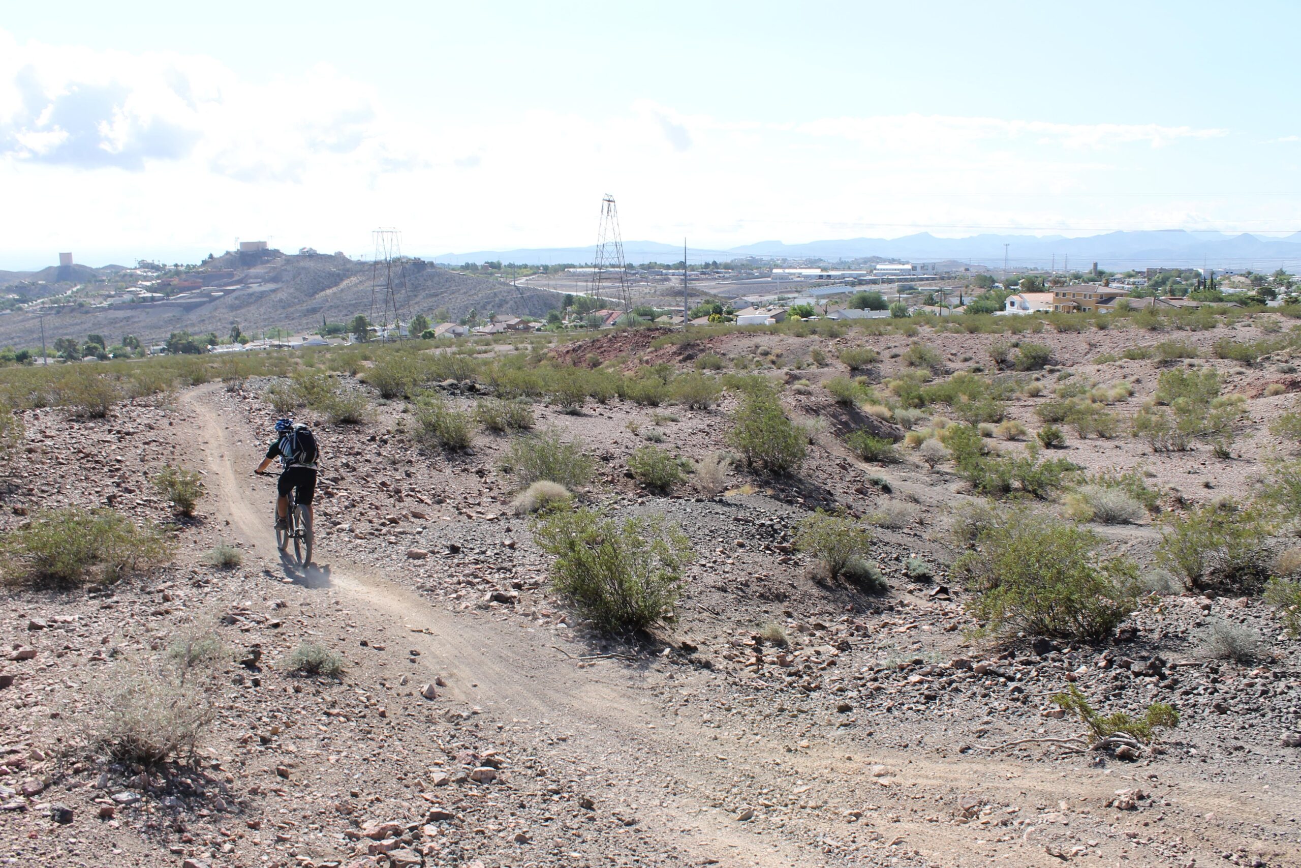 A mountain biker rides along a dirt trail in a rocky desert landscape, with sparse vegetation and distant hills visible in the background. Power lines and buildings can be seen in the distance under a clear sky. Bootleg Canyon mountain bike trail.