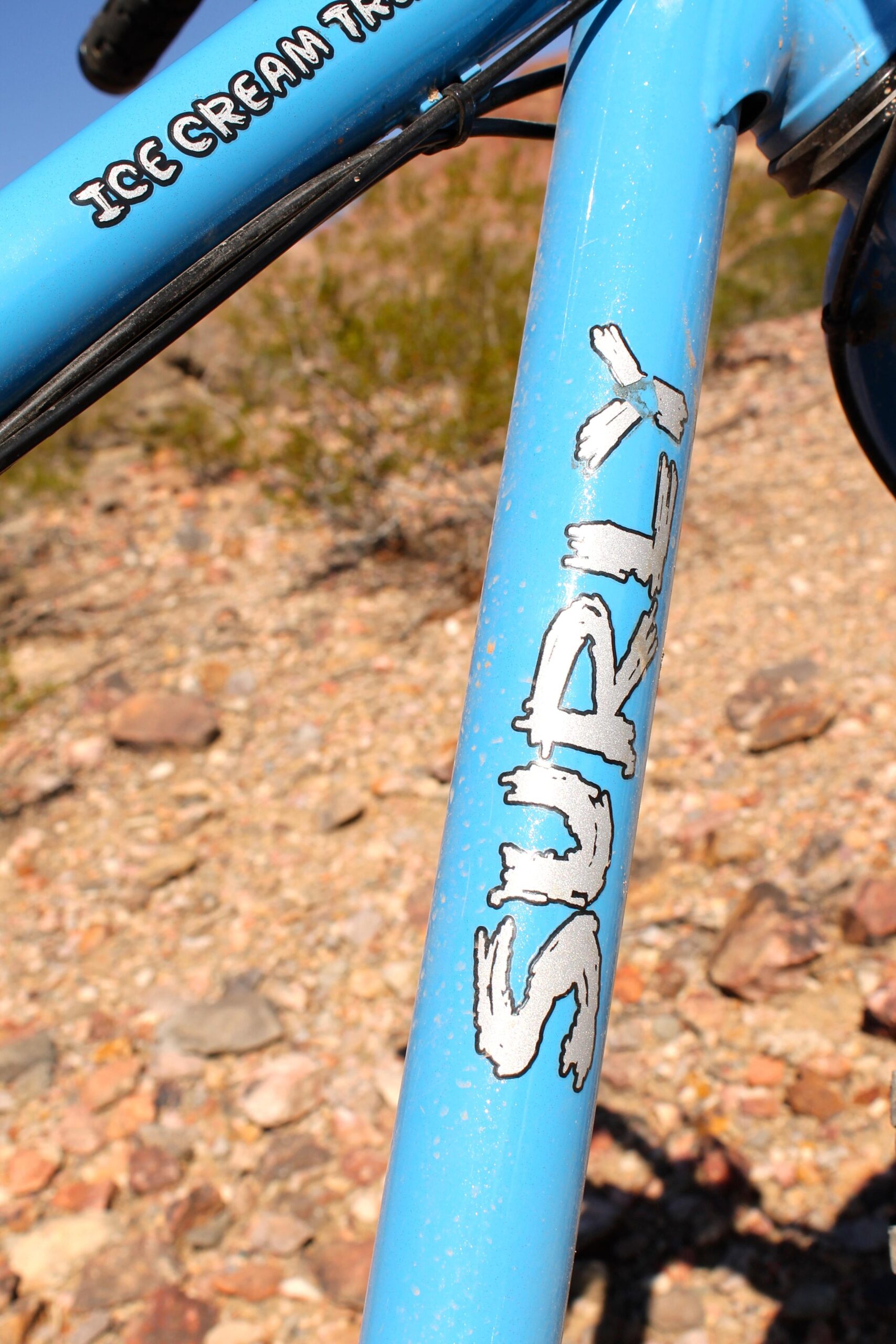 Surly Ice Cream Truck: Close-up of a blue bicycle frame featuring the "Surly" logo and the text "Ice Cream Truck." The frame is situated on a rocky, outdoor terrain with some vegetation in the background.