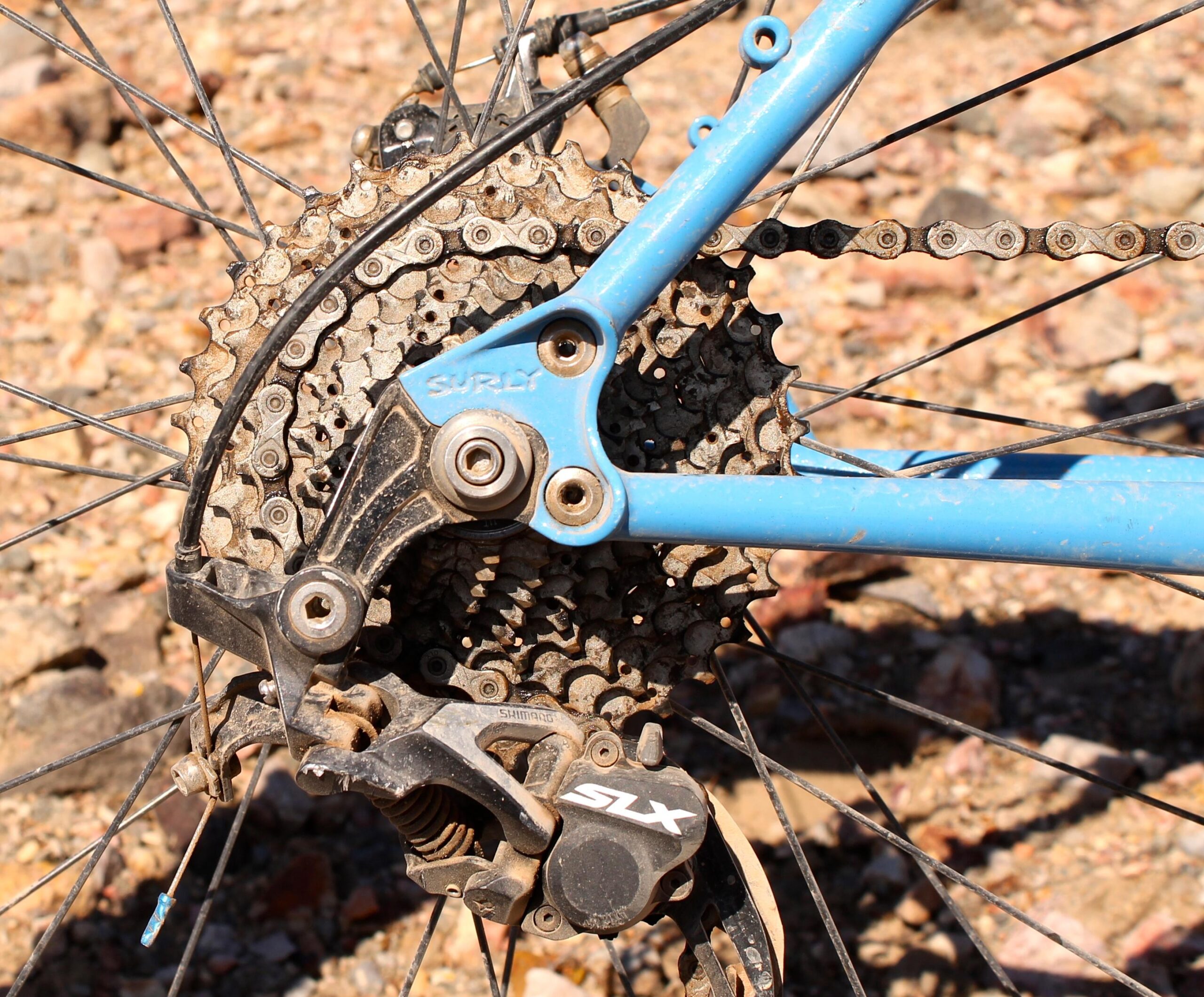 Surly Ice Cream Truck: Close-up of a bicycle's rear derailleur and cassette, featuring a blue frame with a visible trademark logo and a mix of dirt and dust on the components, set against a rocky ground background.