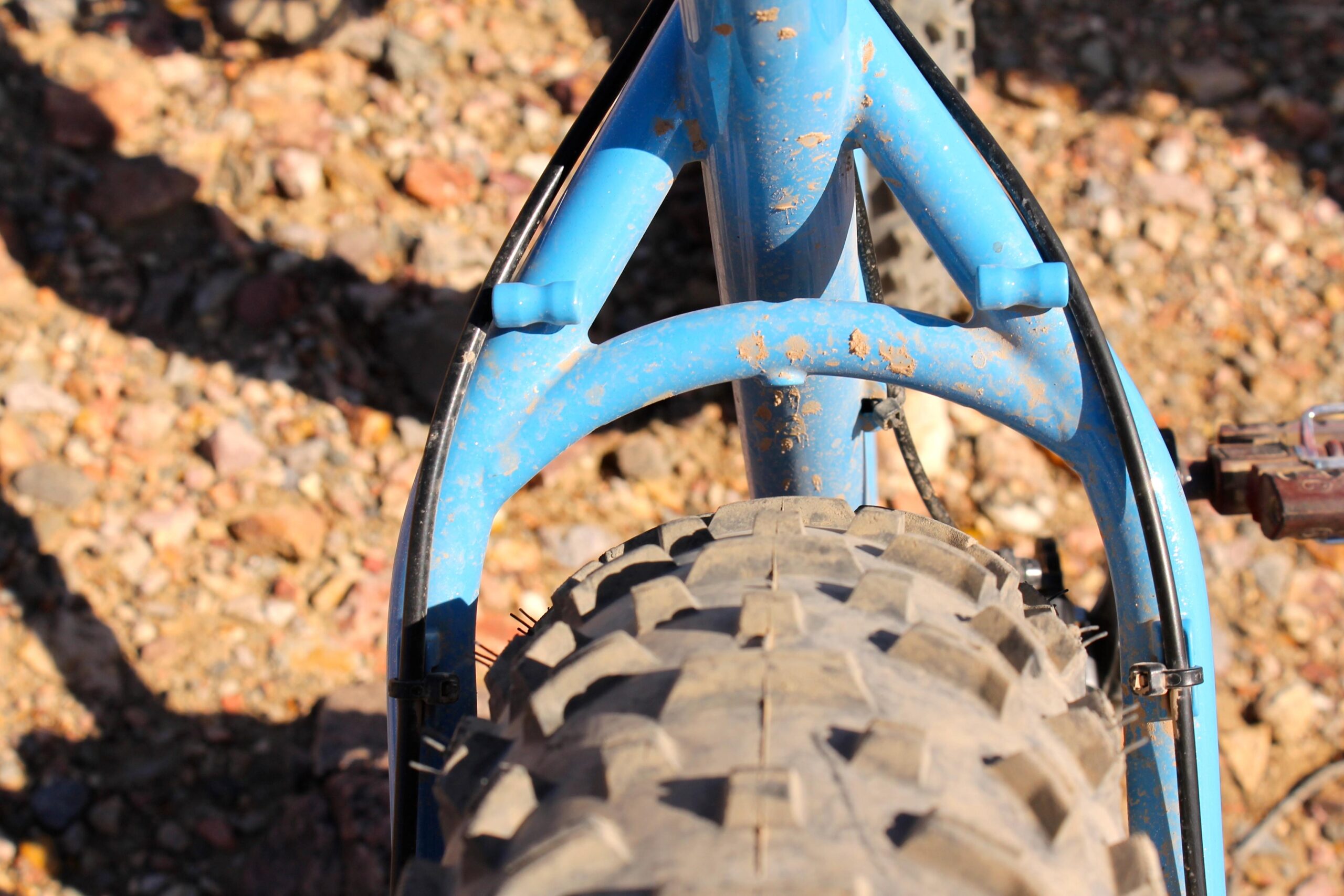 Surly Ice Cream Truck: Close-up view of the rear section of a blue bicycle frame, highlighting the tire and suspension system. The image shows a rugged tire tread and details of the bike's frame, set against a background of gravel and small rocks.