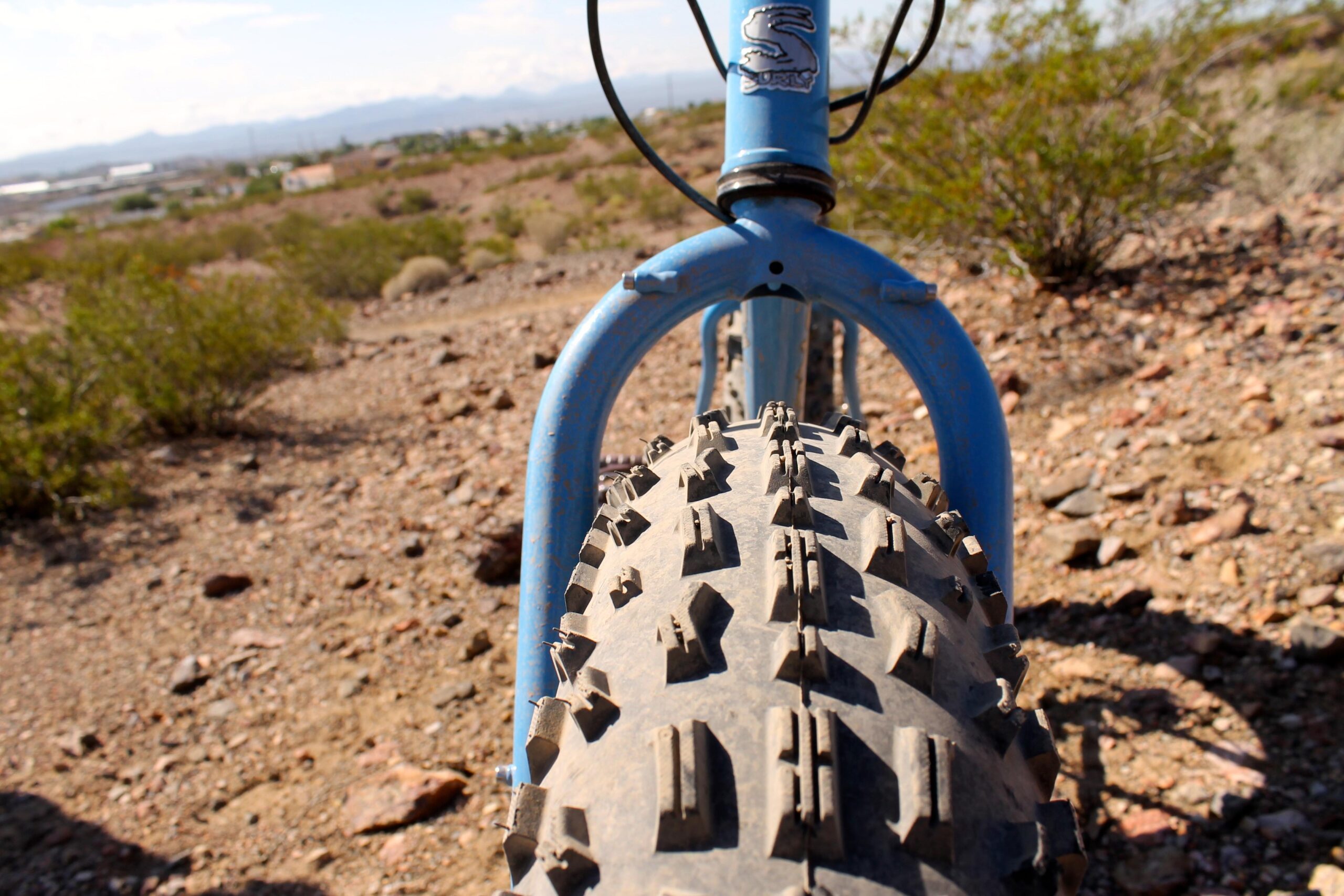 Surly Ice Cream Truck: Close-up view of a blue bicycle frame and tire on rocky terrain, showcasing the rugged tread of the tire designed for off-road biking. In the background, there are sparse desert plants and distant mountains under a clear sky.