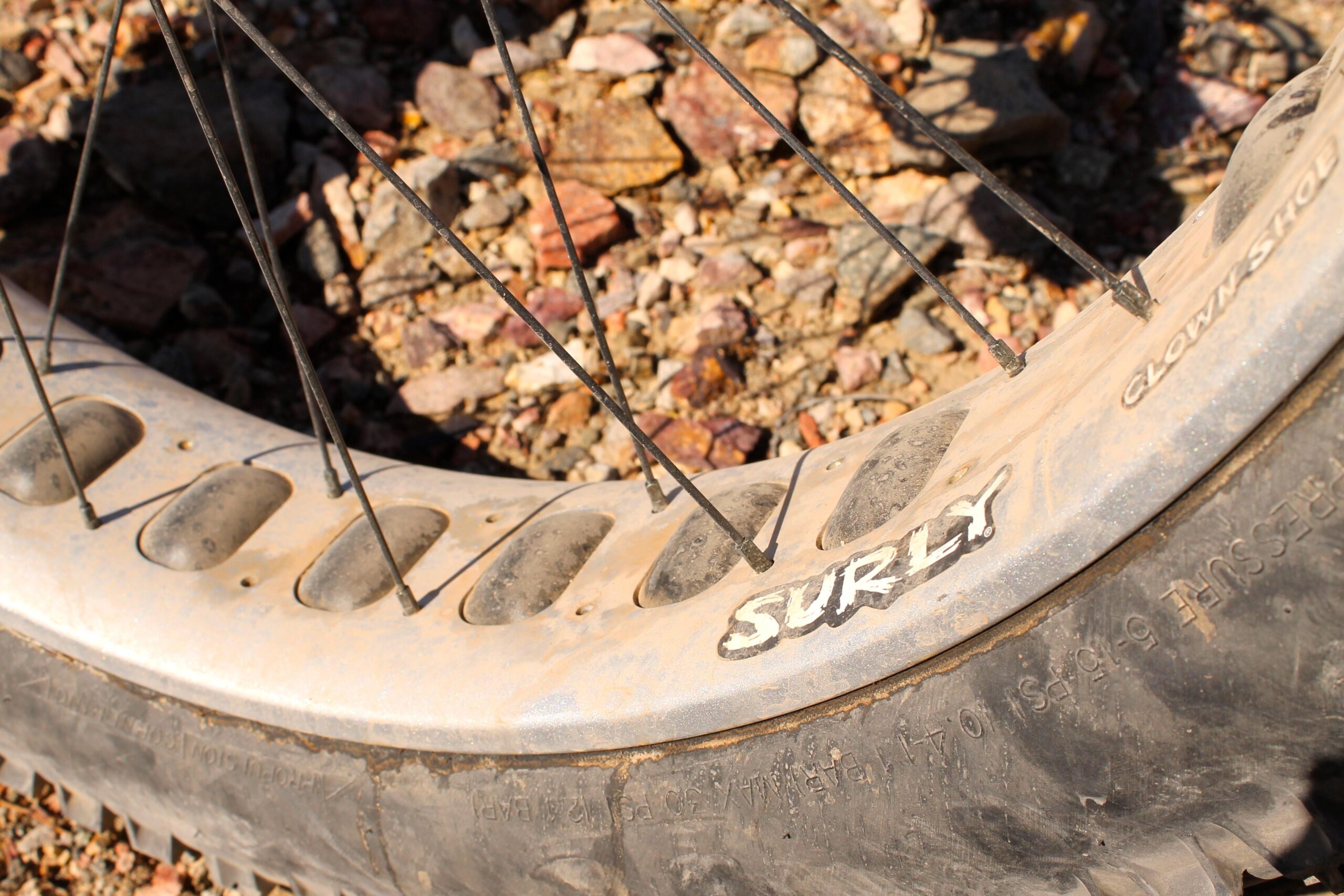 Surly Ice Cream Truck: Close-up view of a dirty bicycle wheel with a fat tire, featuring the brand name "Surly" on the rim. The wheel includes metal spokes and is set against a background of gravel and rocks.