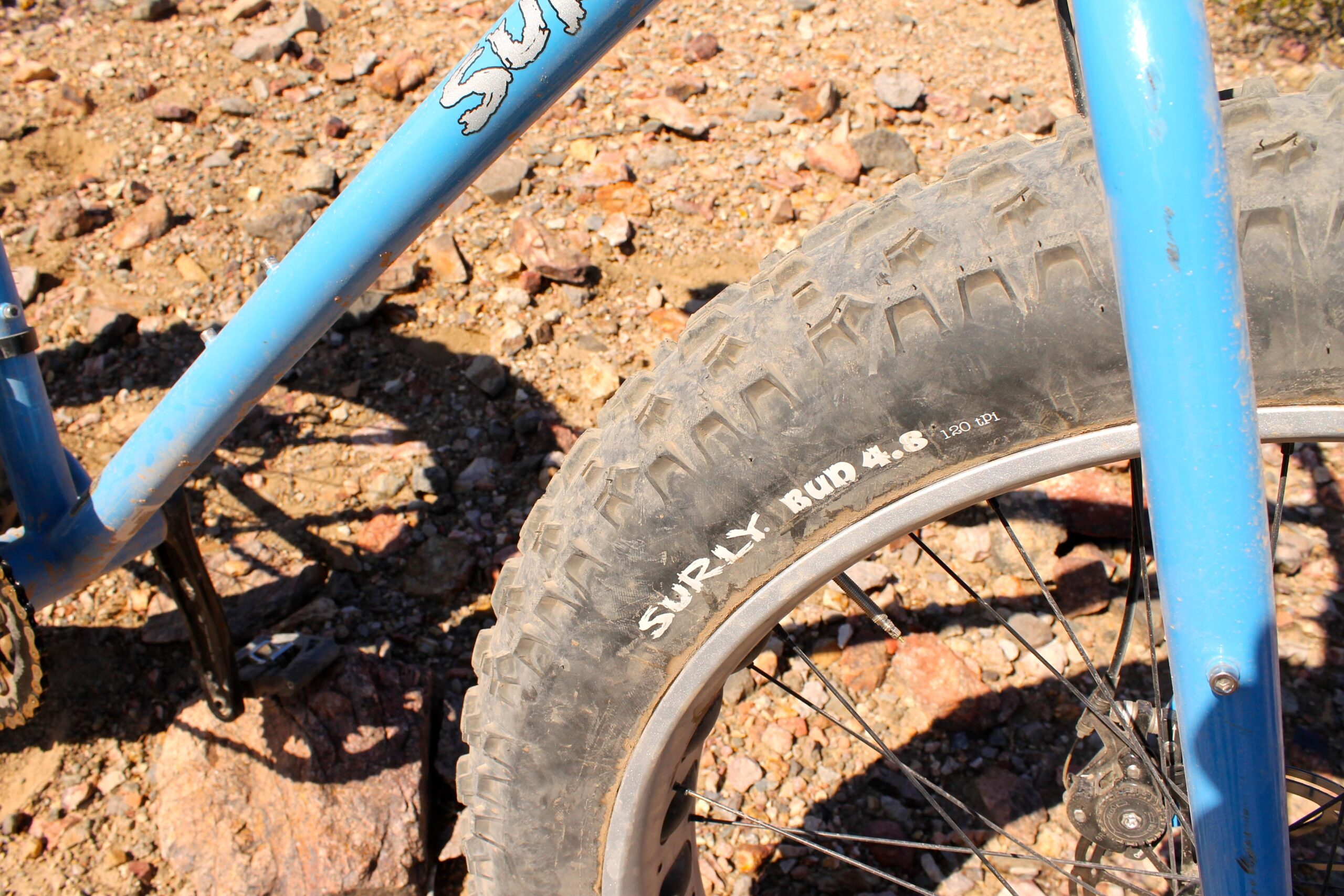 Surly Ice Cream Truck: Close-up view of a blue bicycle frame with a "Surly" logo and a wide, rugged tire labeled "SURLY BUD 4.6," resting on rocky terrain.