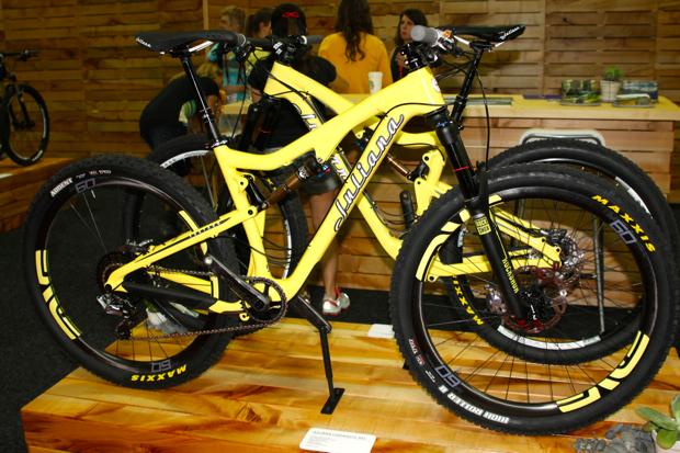 Juliana Furtado: A bright yellow mountain bike is displayed on a wooden platform, showcasing its sleek design and features. The bike has a distinctive frame with noticeable branding and is equipped with thick tires designed for rugged terrain. In the background, several people can be seen engaged in conversation at a booth, surrounded by a wooden backdrop.