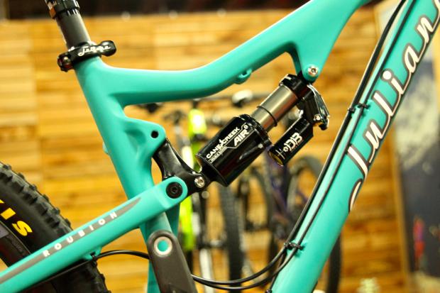Juliana Roubion: Close-up view of a turquoise mountain bike frame featuring a rear shock and suspension components, set against a blurred background of bicycles.