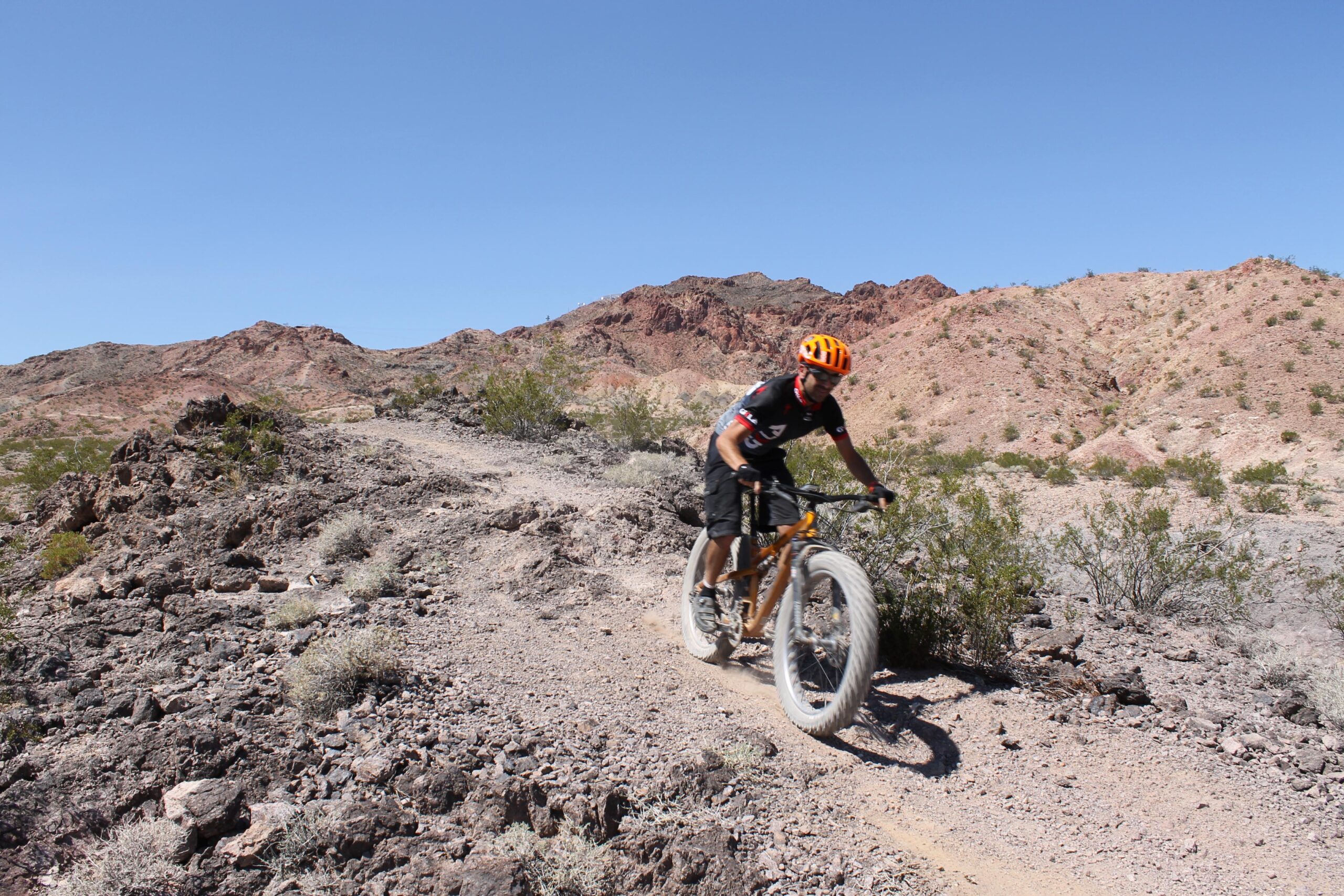 A person riding a mountain bike on a rugged dirt trail in a desert landscape, with rocky hills and sparse vegetation in the background under a clear blue sky. Bootleg Canyon mountain bike trail.