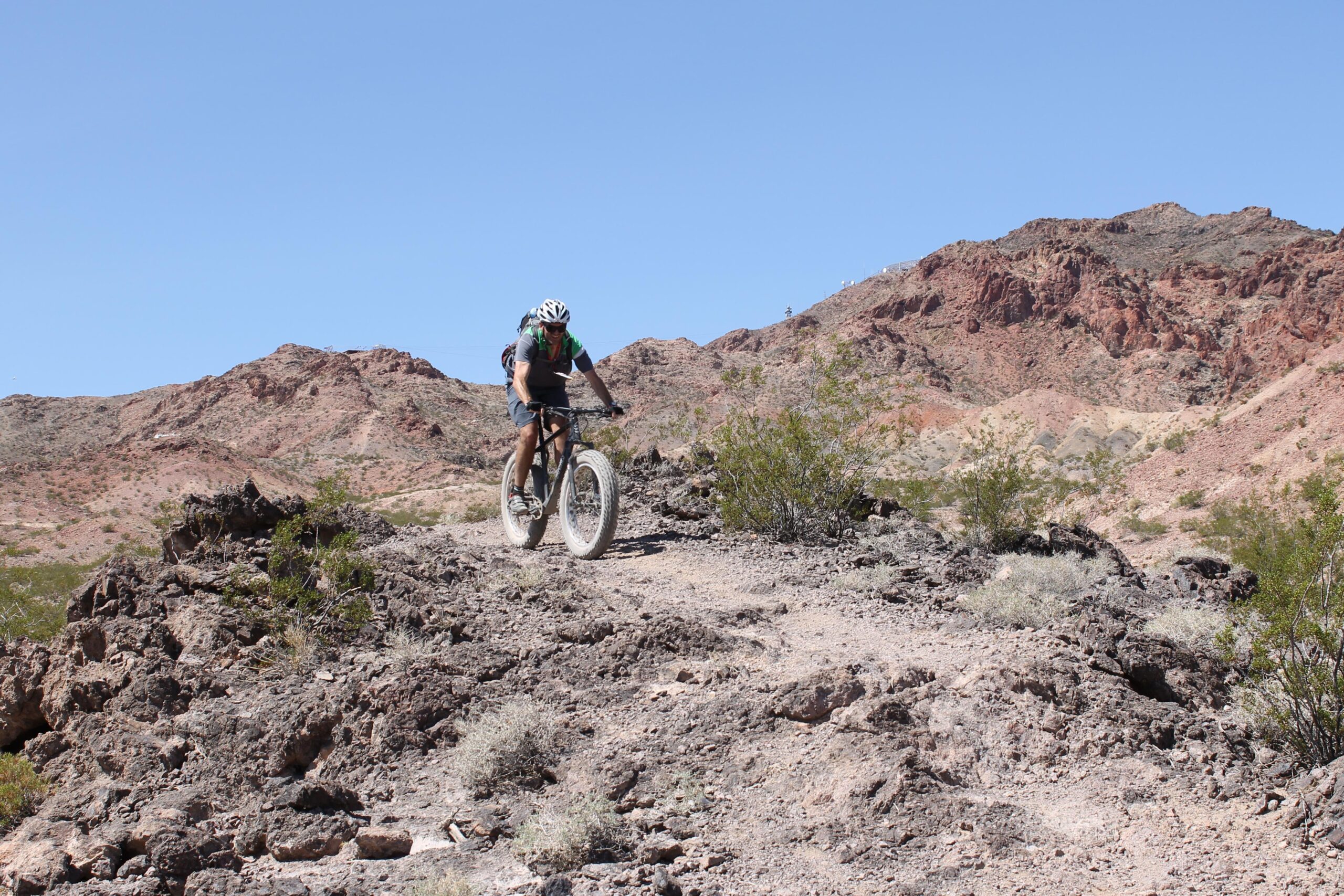 A person riding a fat-tire bicycle on a rocky trail in a desert landscape, with rugged mountains in the background and a clear blue sky above. Bootleg Canyon mountain bike trail.