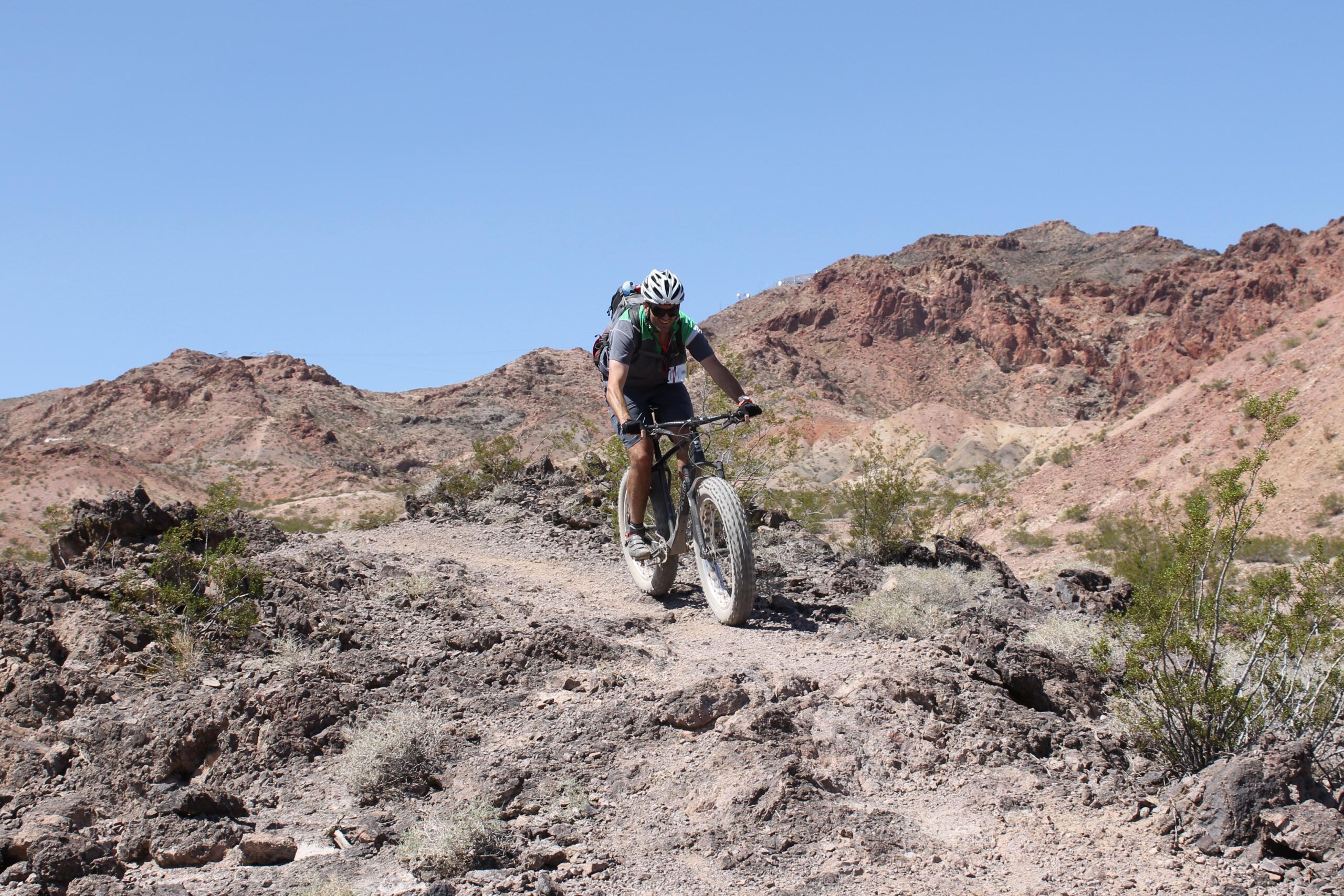 A cyclist riding a fat bike on a rocky trail in a desert landscape, with red mountains and clear blue skies in the background. Bootleg Canyon mountain bike trail.