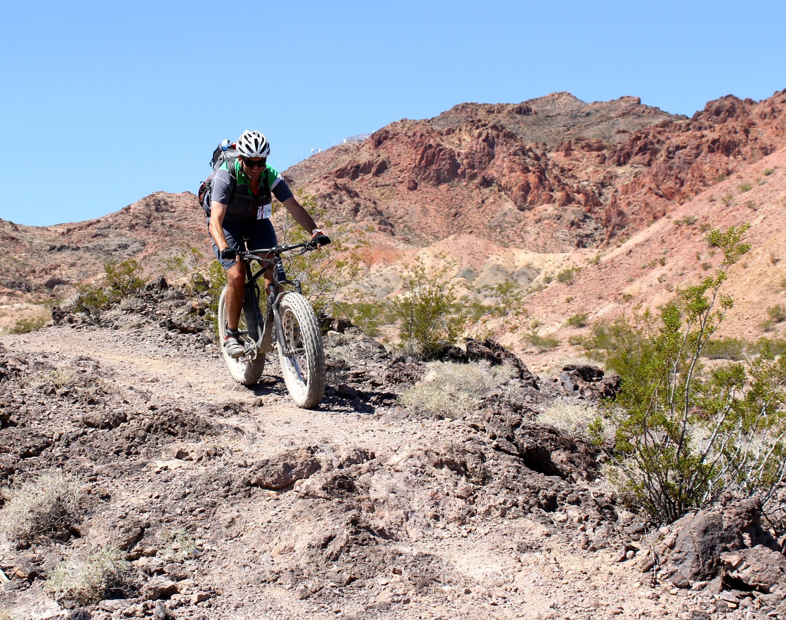 Borealis Echo: A cyclist rides a fat tire bike along a rugged trail in a rocky desert landscape, surrounded by colorful mountains under a clear blue sky. The terrain is uneven with patches of vegetation, showcasing a challenging outdoor adventure.