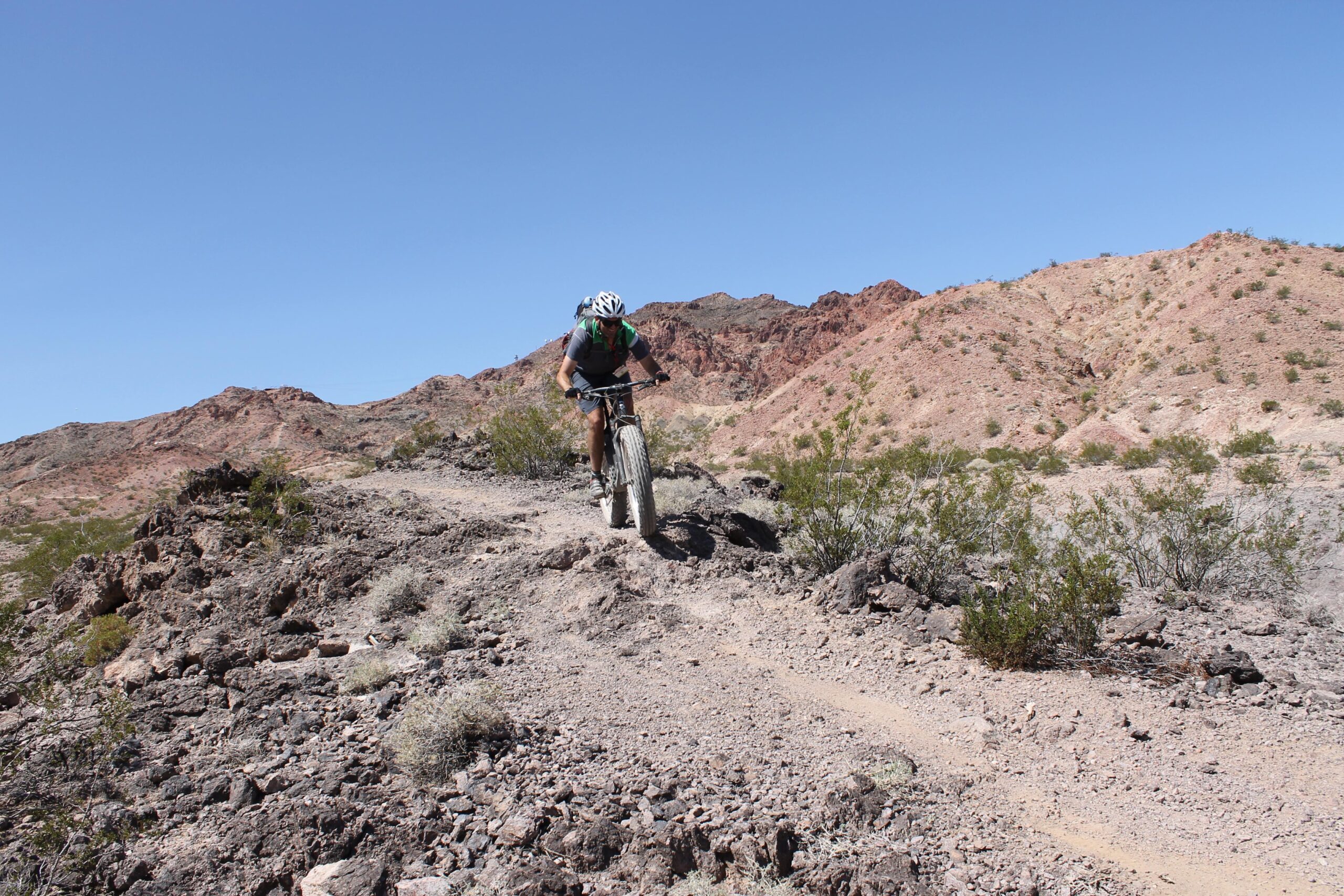 A mountain biker navigating a rocky trail in a desert landscape, surrounded by rolling hills and sparse vegetation under a clear blue sky. Bootleg Canyon mountain bike trail.