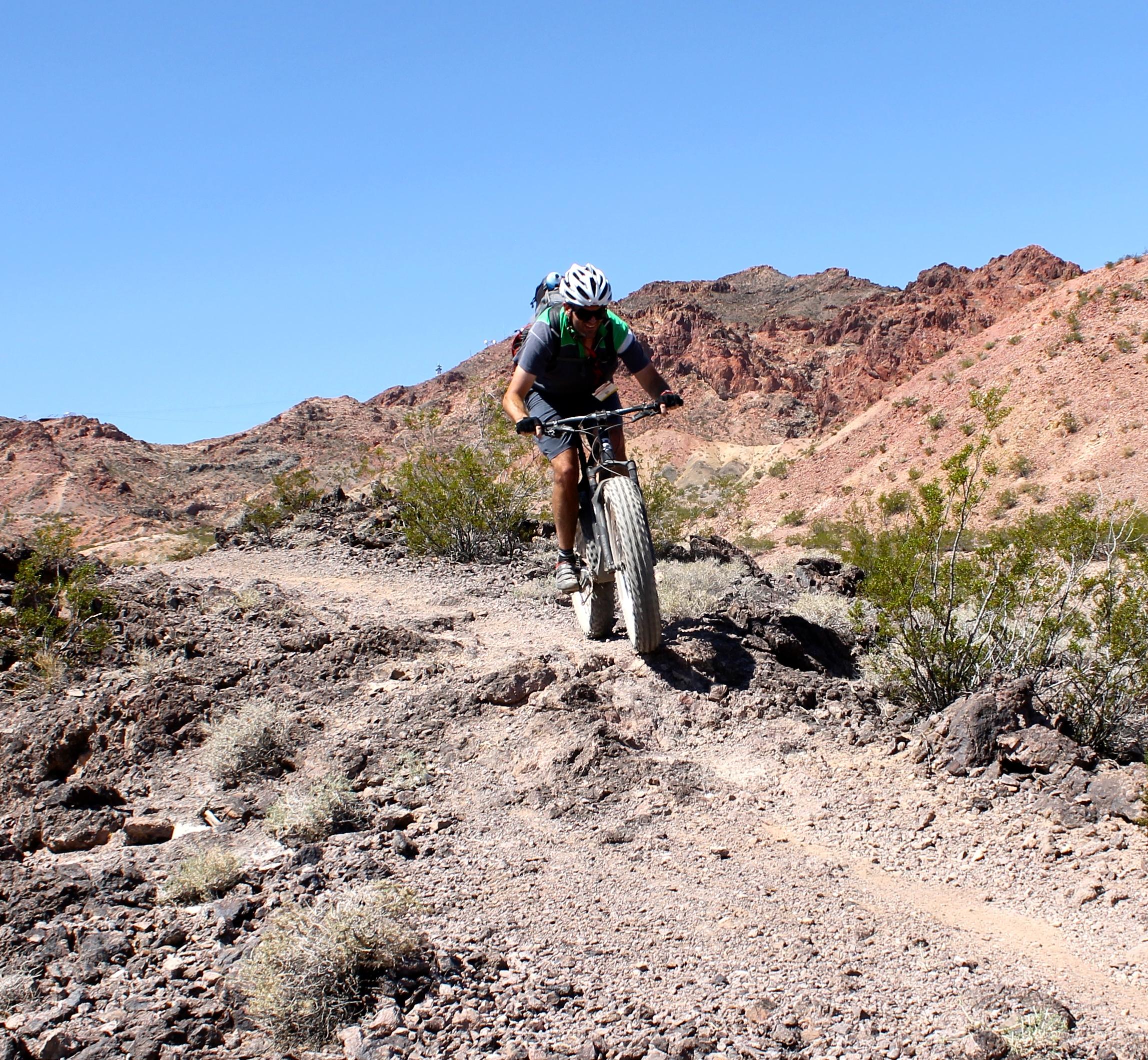 Borealis Echo: A mountain biker navigating a rocky trail in a barren, mountainous landscape under a clear blue sky. Vegetation and rocky terrain surround the path, showcasing a rugged outdoor setting.