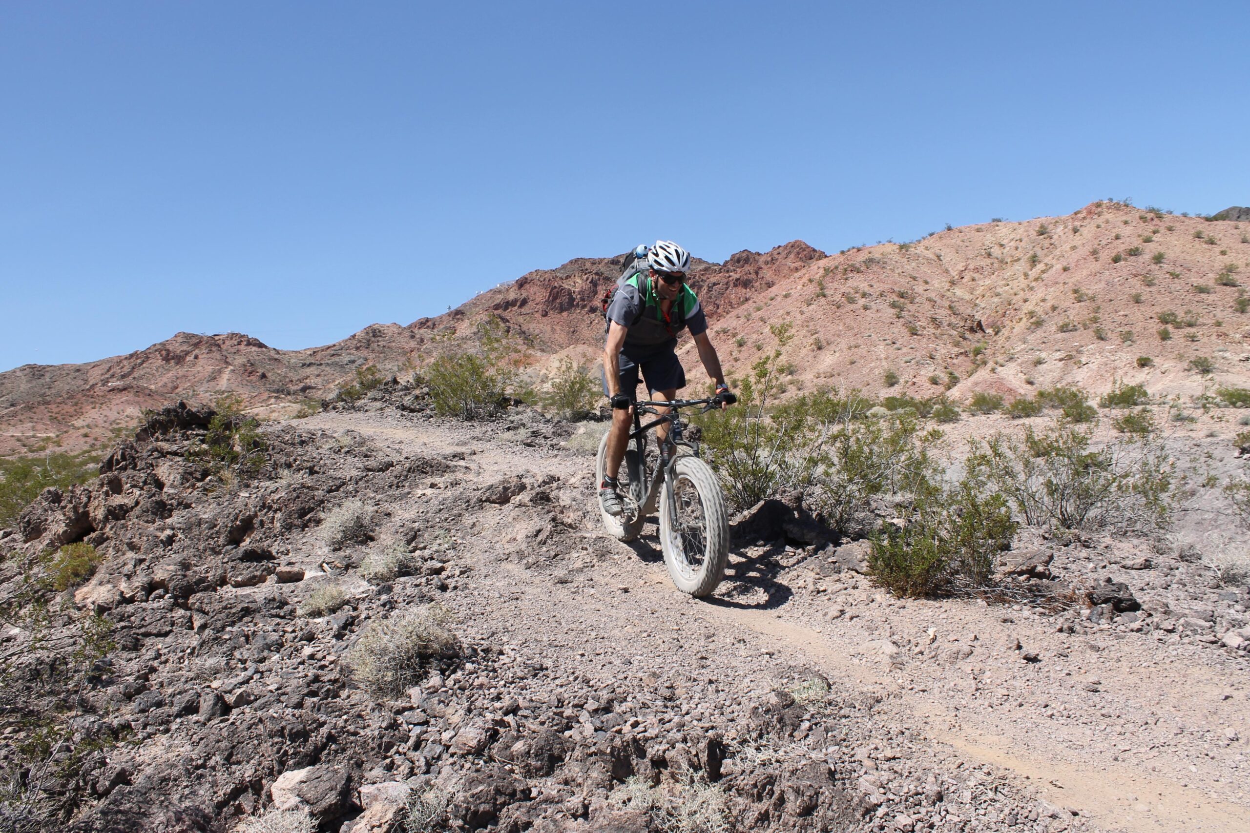 A mountain biker navigating a rocky trail in a desert landscape, surrounded by red and brown hills under a clear blue sky. Bootleg Canyon mountain bike trail.