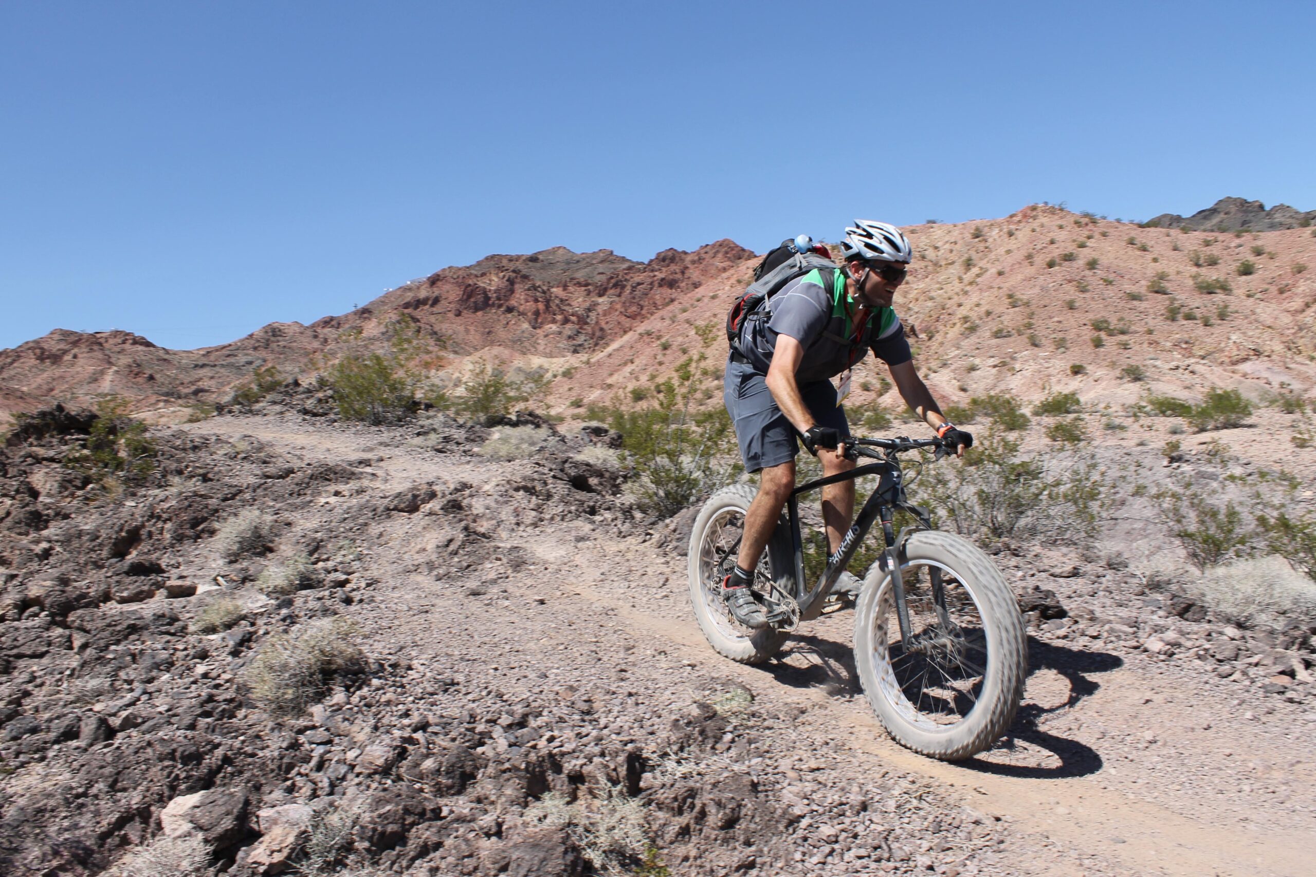 A person riding a mountain bike on a rocky trail in a desert landscape, surrounded by hills and sparse vegetation under a clear blue sky. The biker wears a helmet and a backpack. Bootleg Canyon mountain bike trail.