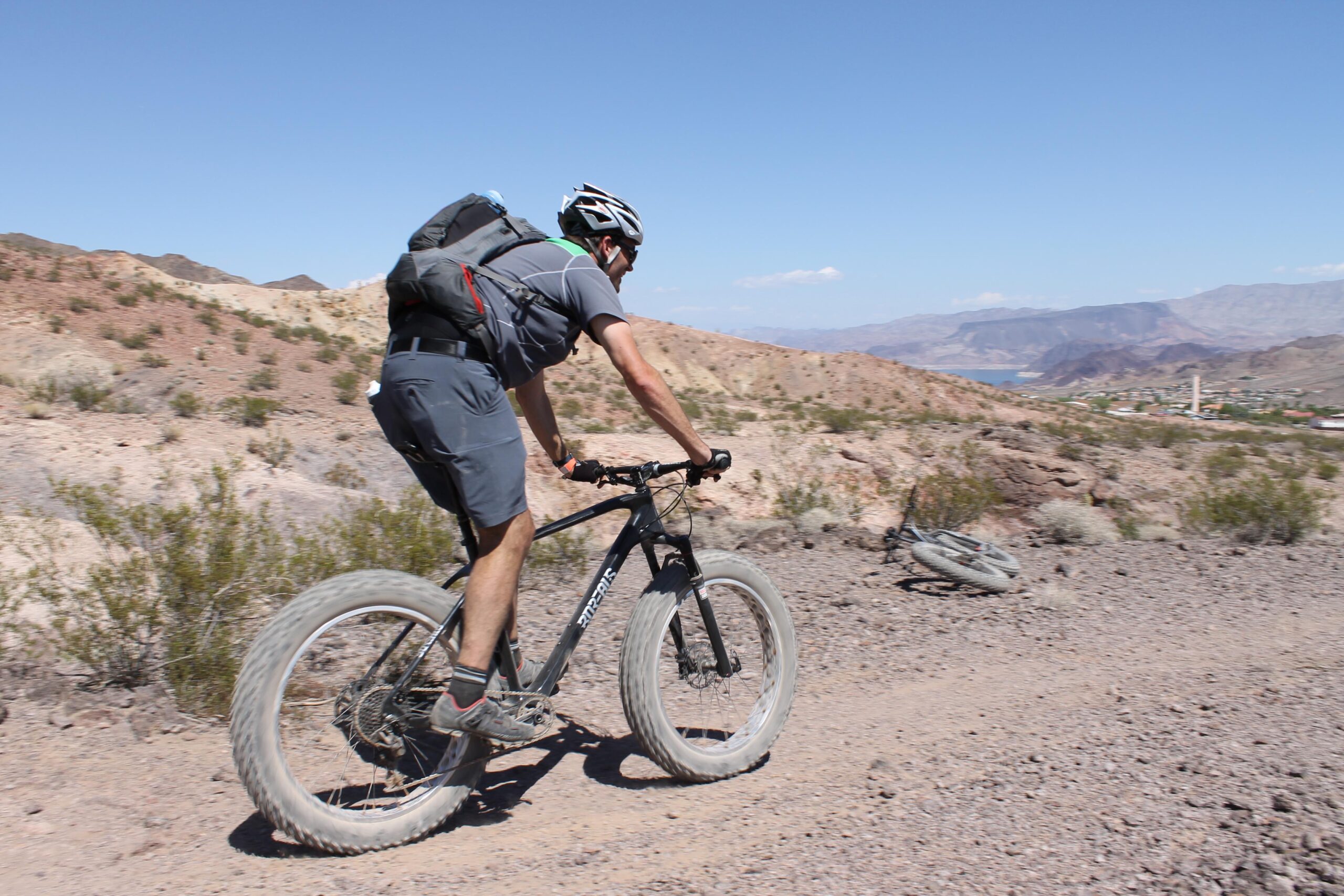 A person riding a mountain bike on a rocky trail, surrounded by desert terrain and mountains in the background. The cyclist is wearing a helmet and a backpack, with a focus on the bike's large tires. A distant view of a landscape with a lake and small buildings is visible on the horizon under a clear blue sky. Bootleg Canyon mountain bike trail.