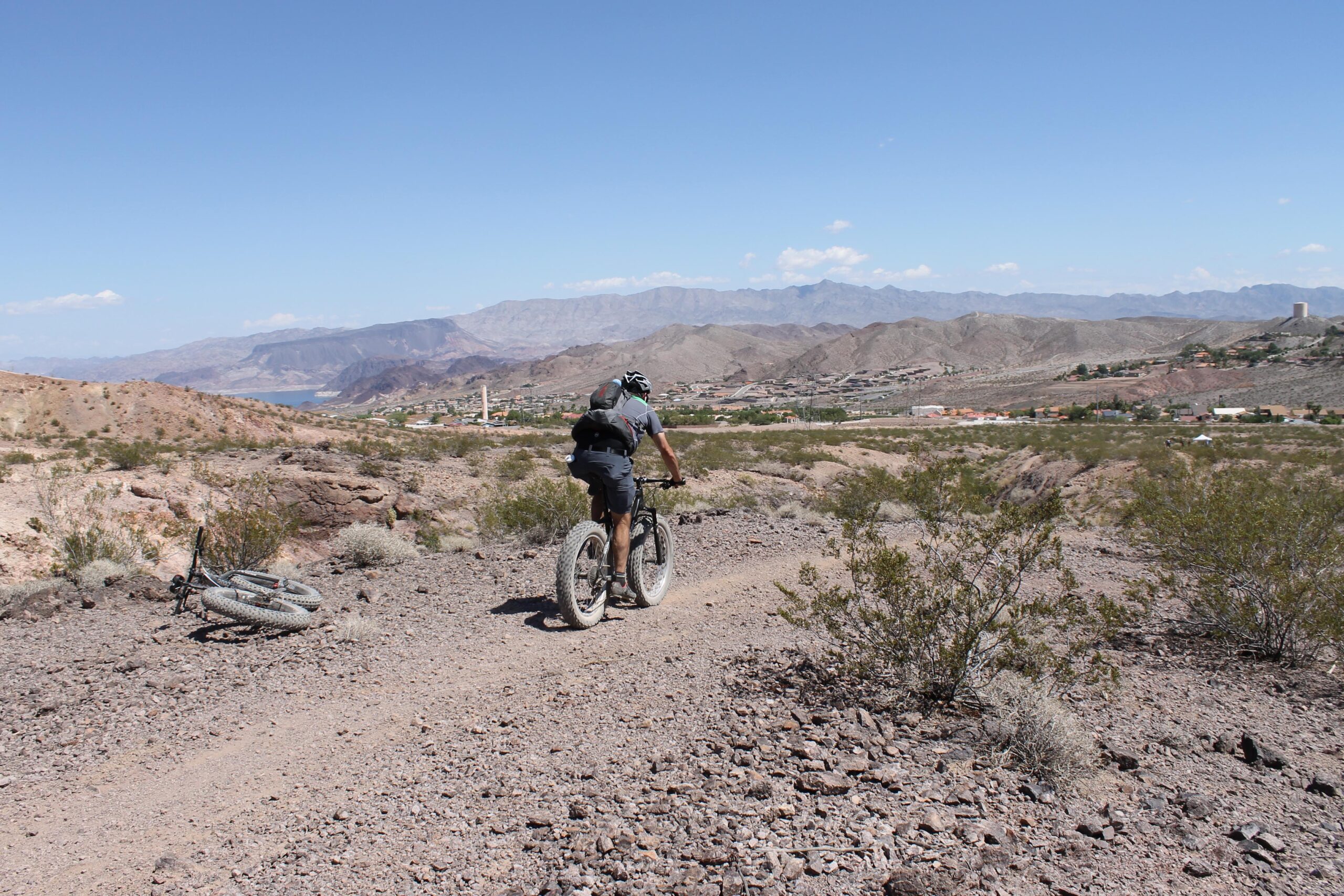 A person riding a fat bike along a dirt trail in a rocky desert landscape, with mountains and a lake visible in the background under a clear blue sky. A second bike lies on the ground nearby. Bootleg Canyon mountain bike trail.