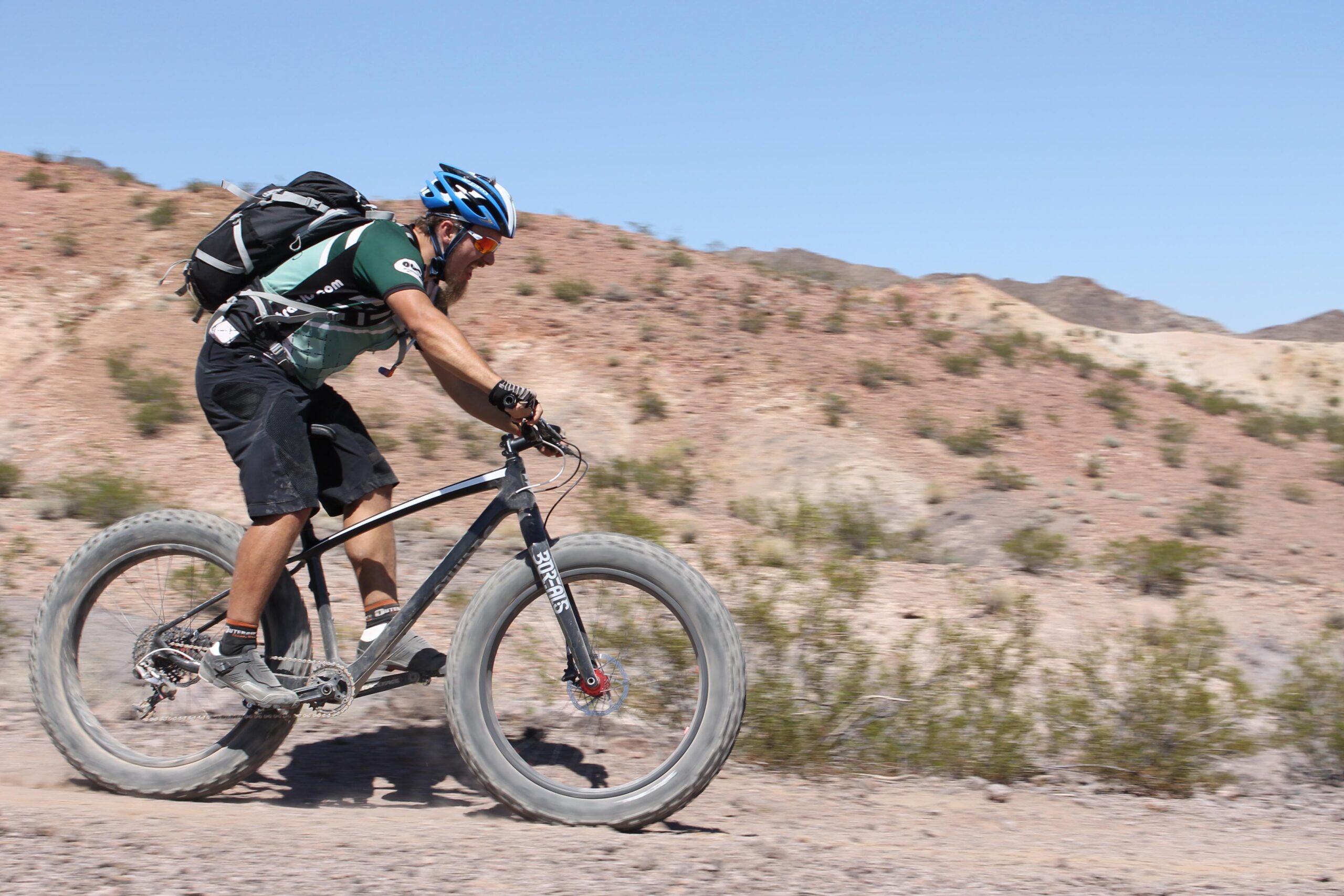 A cyclist dressed in a green and black jersey rides a black fat bike along a dusty trail in a desert landscape, with rocky hills and sparse vegetation in the background. The rider wears a blue helmet and sunglasses, and is depicted in motion, creating a sense of speed and adventure. Bootleg Canyon mountain bike trail.