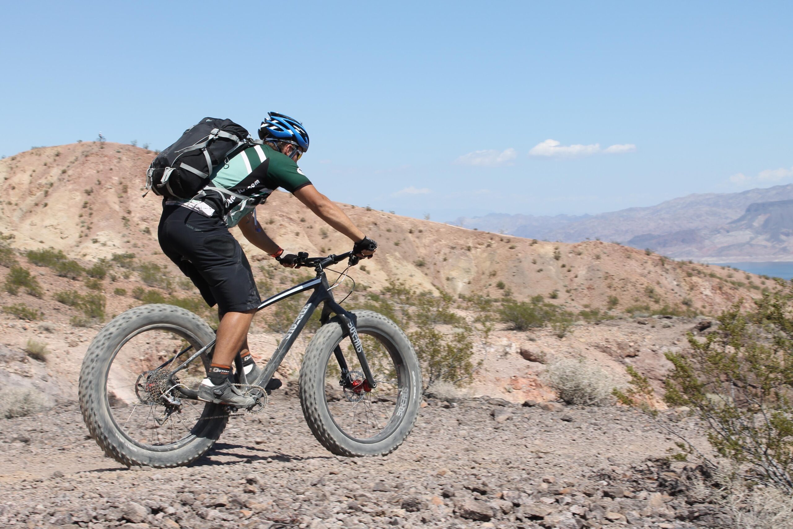 A person riding a fat tire mountain bike on a rocky trail, with hills and distant mountains in the background under a clear blue sky. The rider is wearing a helmet and a backpack, focusing on navigating the terrain. Bootleg Canyon mountain bike trail.