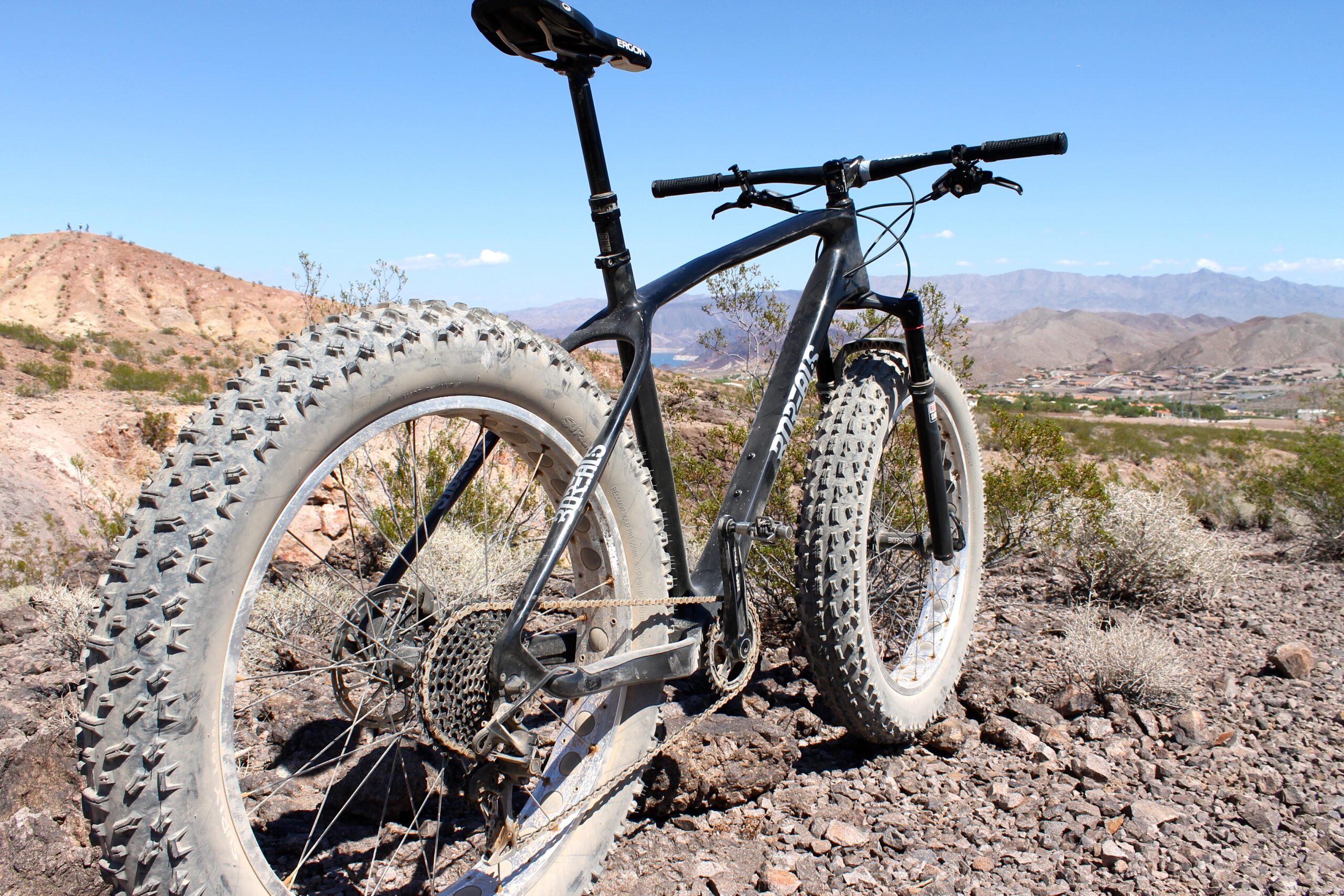 Borealis Echo: A close-up view of a fat tire mountain bike positioned on rocky terrain, showcasing its large, knobby tires and sturdy frame. The background features a desert landscape with hills and distant mountains under a clear blue sky.