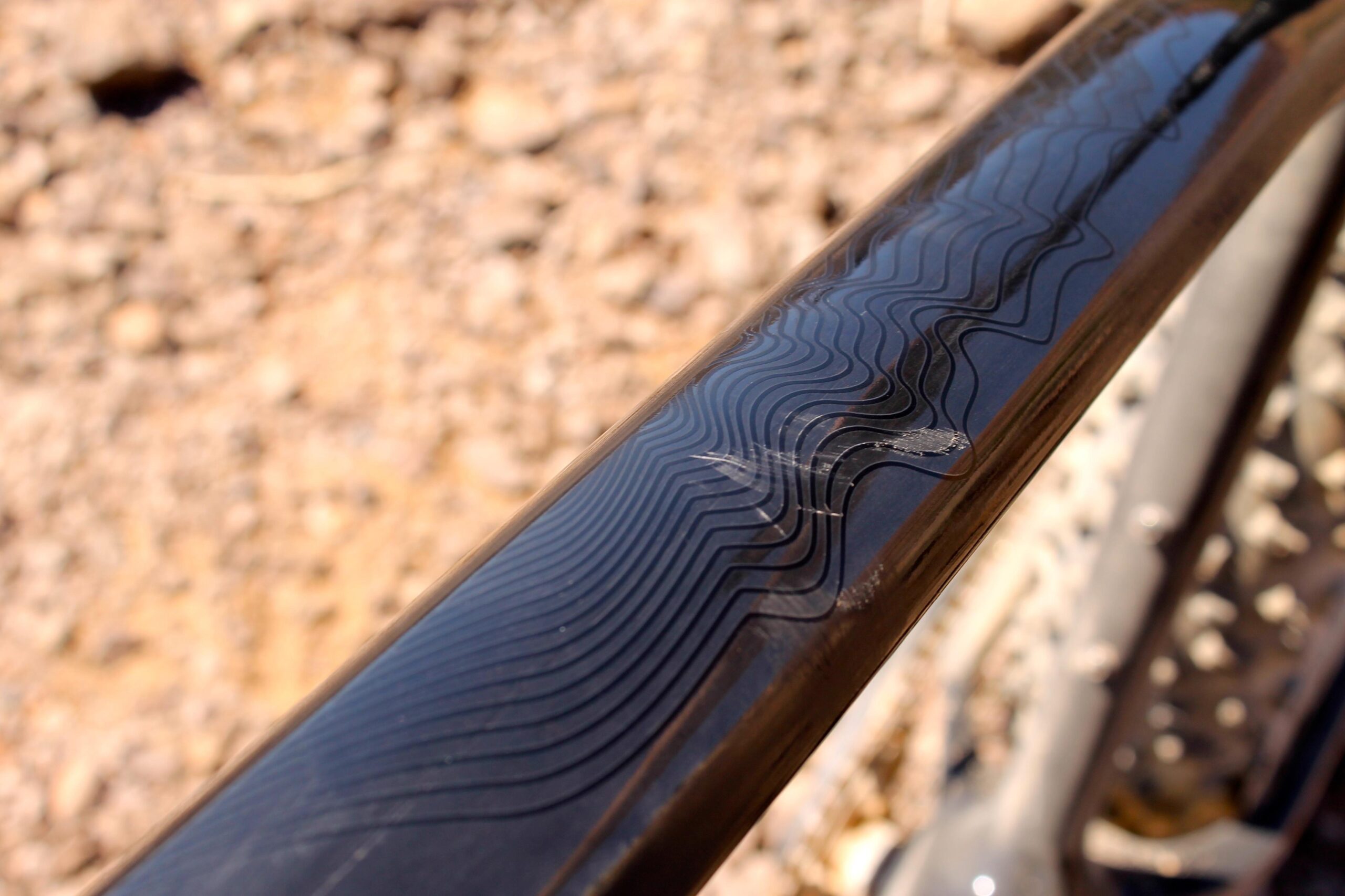 Borealis Echo: Close-up view of a bicycle frame showing a shiny surface with wavy patterns and a small scratch. The background features a blurred, rocky terrain.