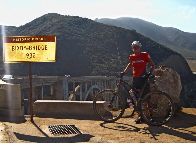 A cyclist in a red jersey stands next to a sign that reads "Historic Bridge Bixby Bridge 1932." The background features a scenic view of rolling green hills and a bridge spanning a canyon. The image captures a sunny day with clear blue skies. The cyclist