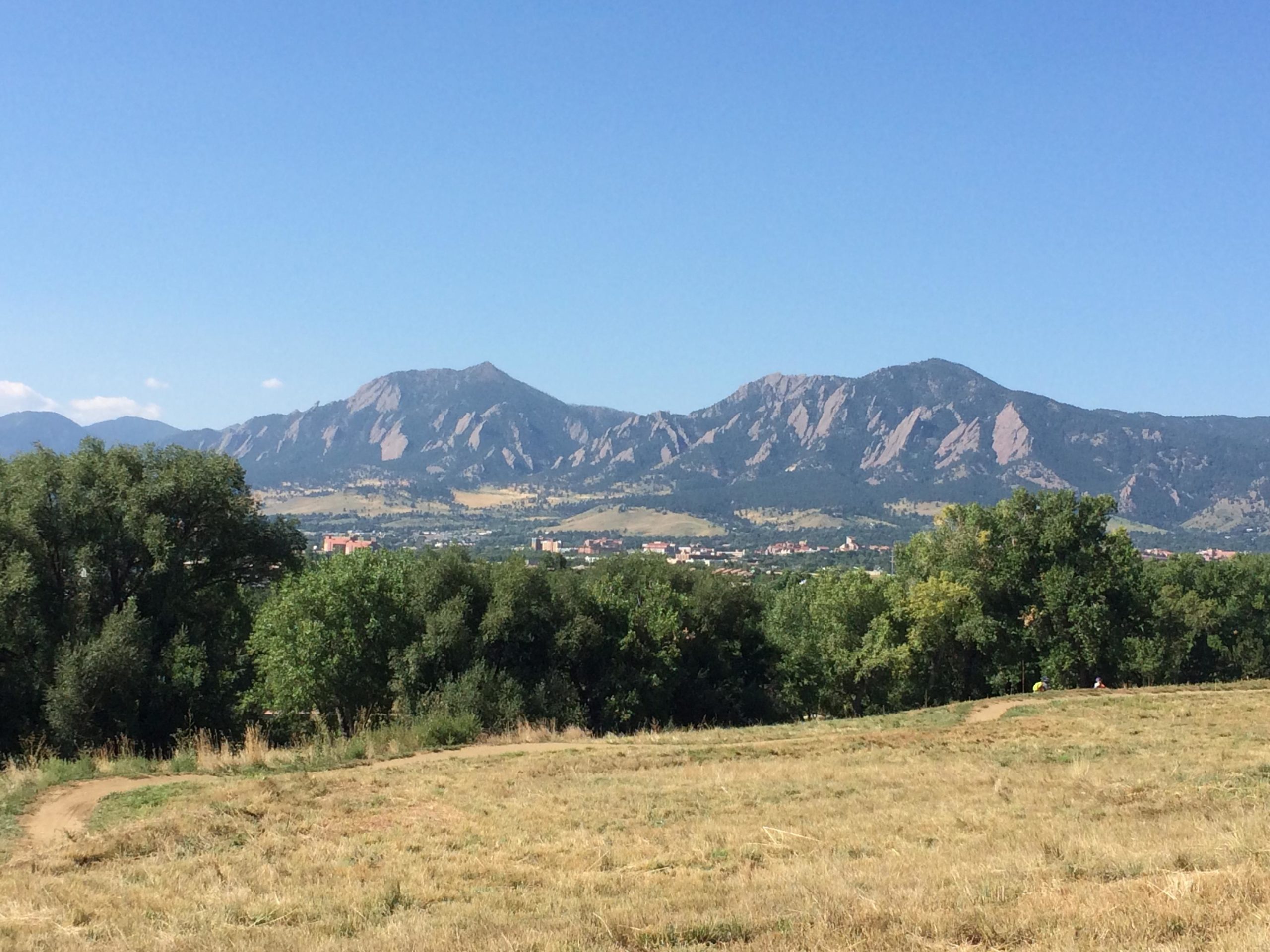 A scenic view of the Flatirons mountain range under a clear blue sky, surrounded by green trees and grassy fields. In the foreground, a path meanders through the landscape, with a glimpse of a small town visible in the valley below. Valmont Bike Park mountain bike trail.