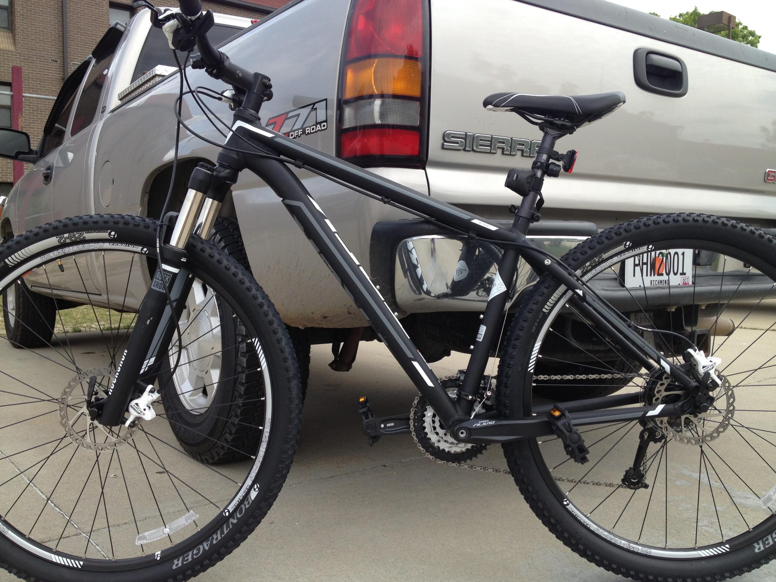 Trek Mamba: A black mountain bike parked next to a silver pickup truck, showcasing the bike's front wheel, suspension fork, and part of the frame against a concrete background.