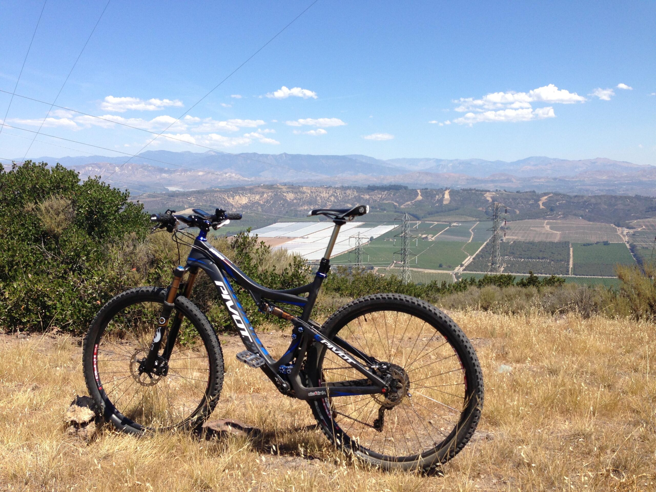 Pivot Mach 429 Carbon: A mountain bike leaning against a rock in a grassy area, with a panoramic view of a valley, fields, and distant mountains under a clear blue sky with a few clouds.