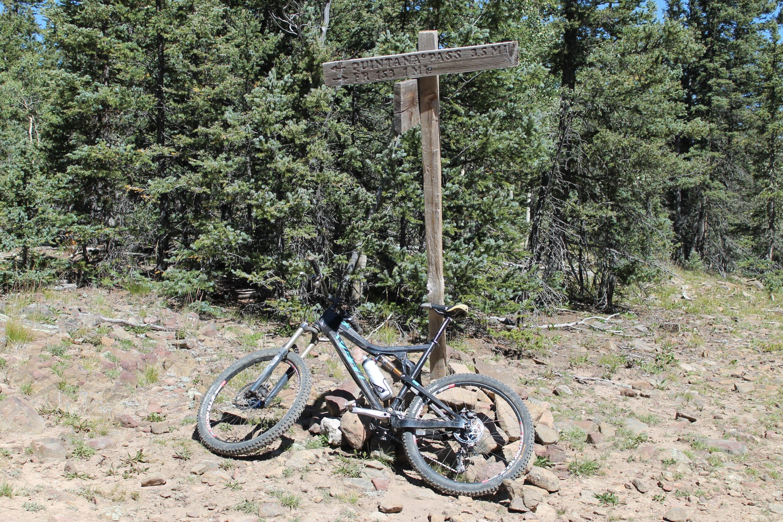 A mountain bike leaned against a wooden trail sign in a forested area, with rocky terrain and greenery in the background. The sign indicates trail distances, with "Qunitana" and "5 miles" visible. South Boundary (164) mountain bike trail.