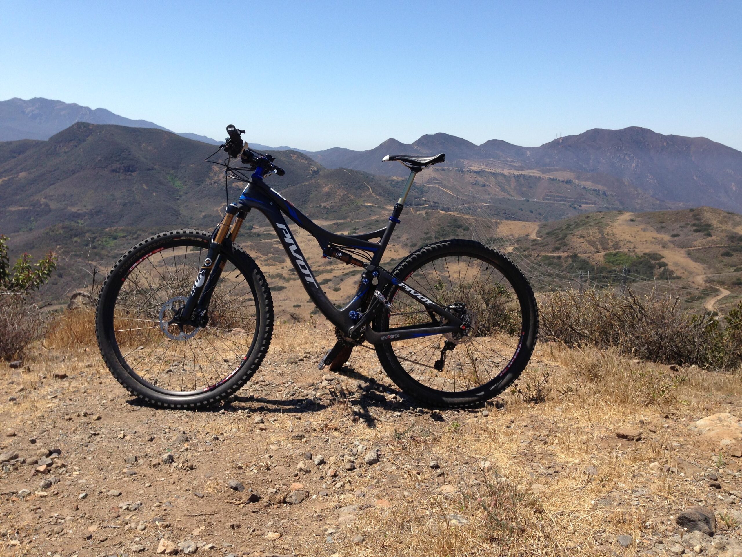 Pivot Mach 429 Carbon: A mountain bike on a rocky outcrop with a view of rolling hills and mountains in the background under a clear blue sky.