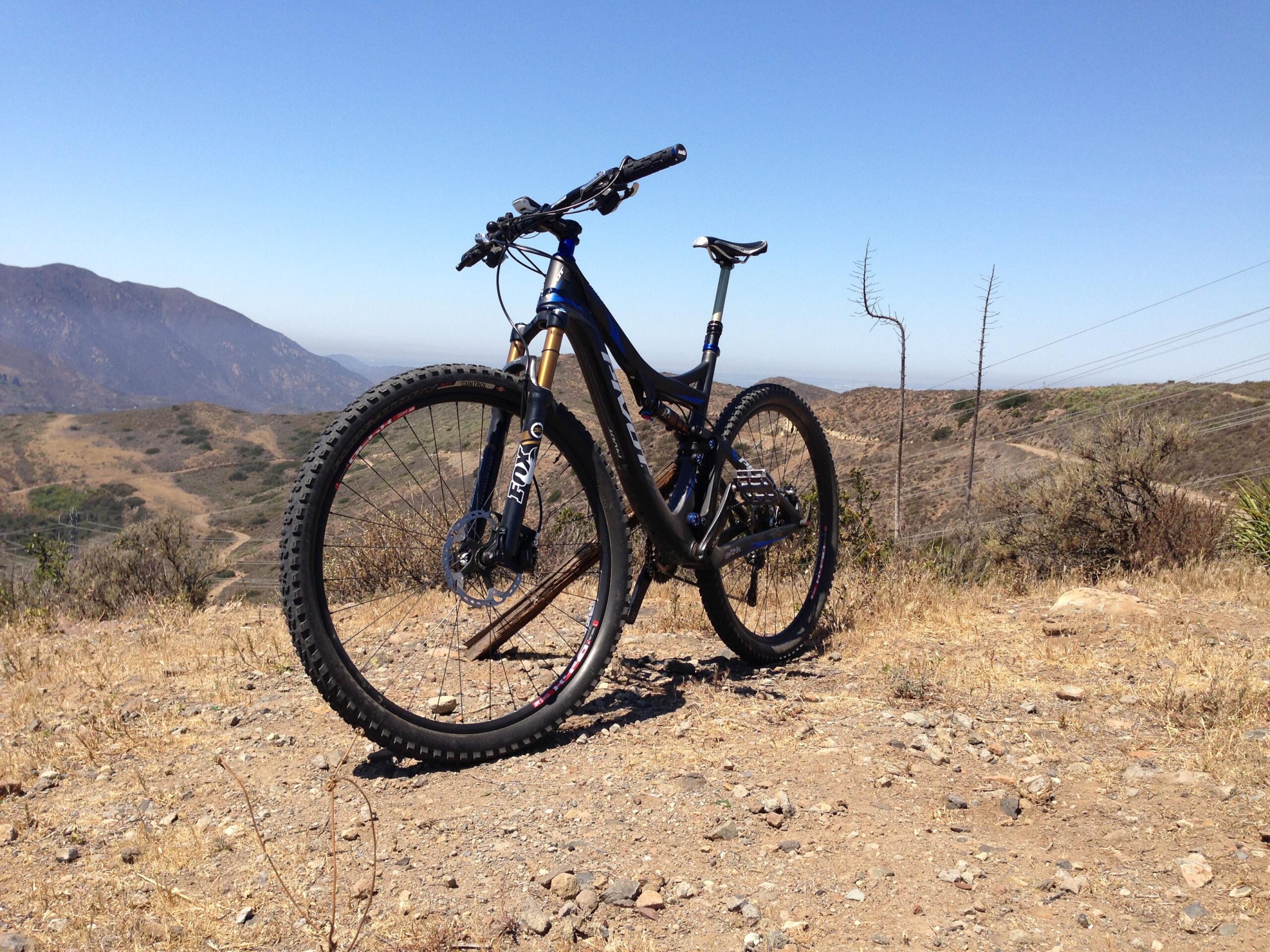 Pivot Mach 429 Carbon: A mountain bike positioned on a rocky, dirt trail with a scenic view of rolling hills and mountains in the background under a clear blue sky.