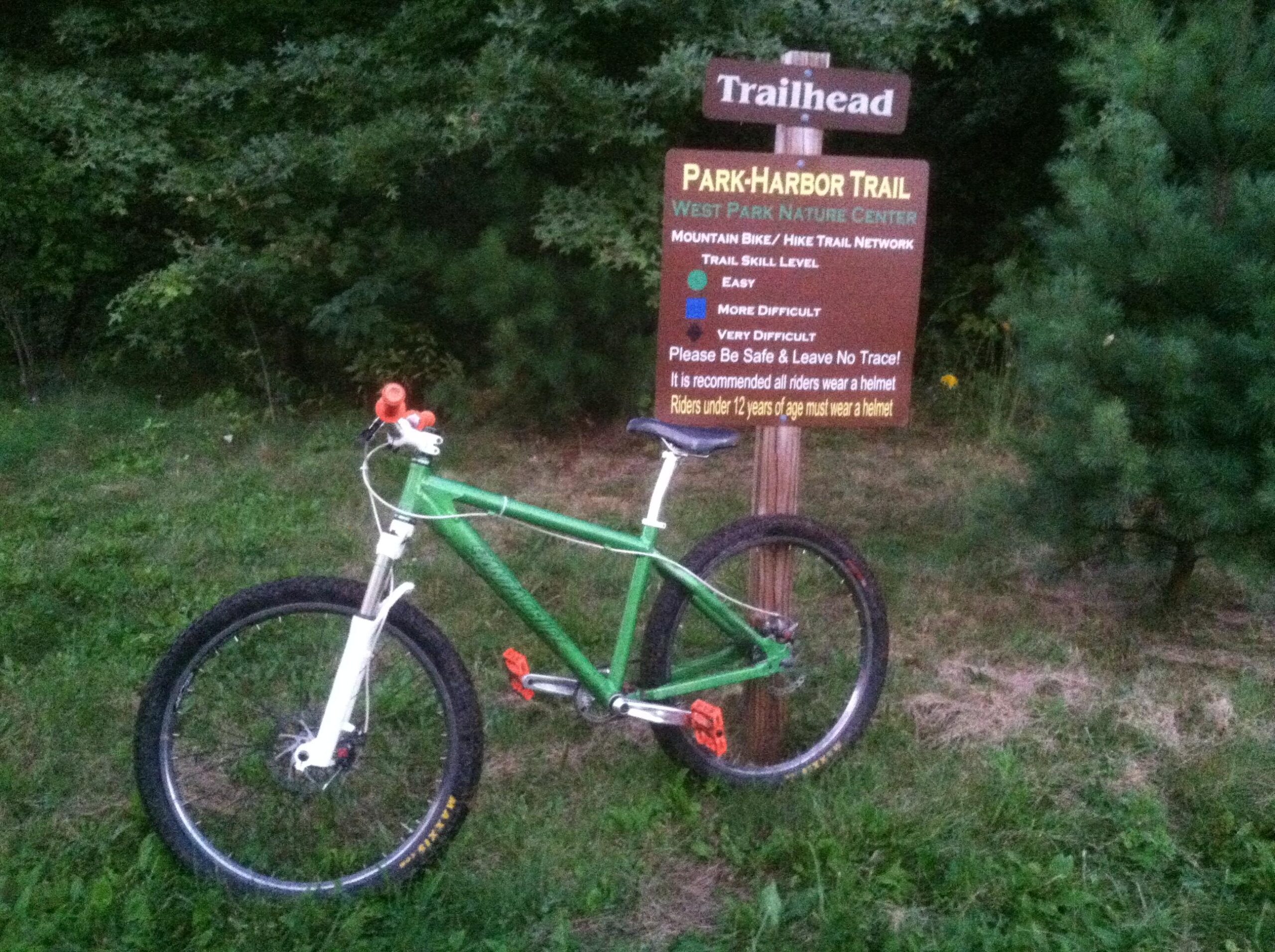 Santa Cruz Chameleon: A green mountain bike parked next to a trailhead sign for the Park-Harbor Trail at West Park Nature Center, indicating trail skill levels (Easy, More Difficult, Very Difficult) and safety recommendations for riders. The background features trees and grass.