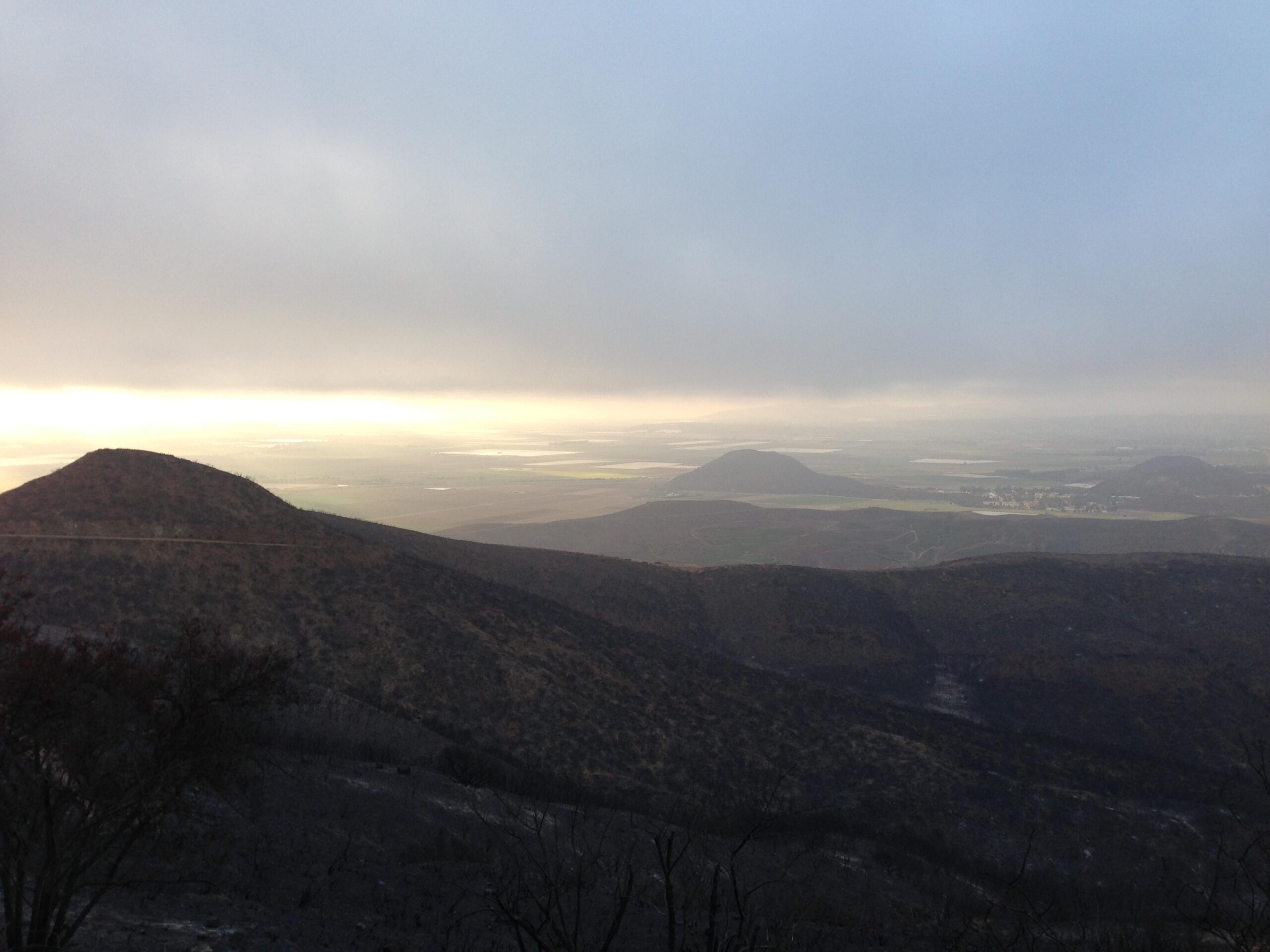 A panoramic view of rolling hills under a cloudy sky, with a faint golden light illuminating the distant landscape. The foreground features dark, textured hills, while the background showcases lush fields and valleys extending towards the horizon. Point Mugu State Park mountain bike trail.