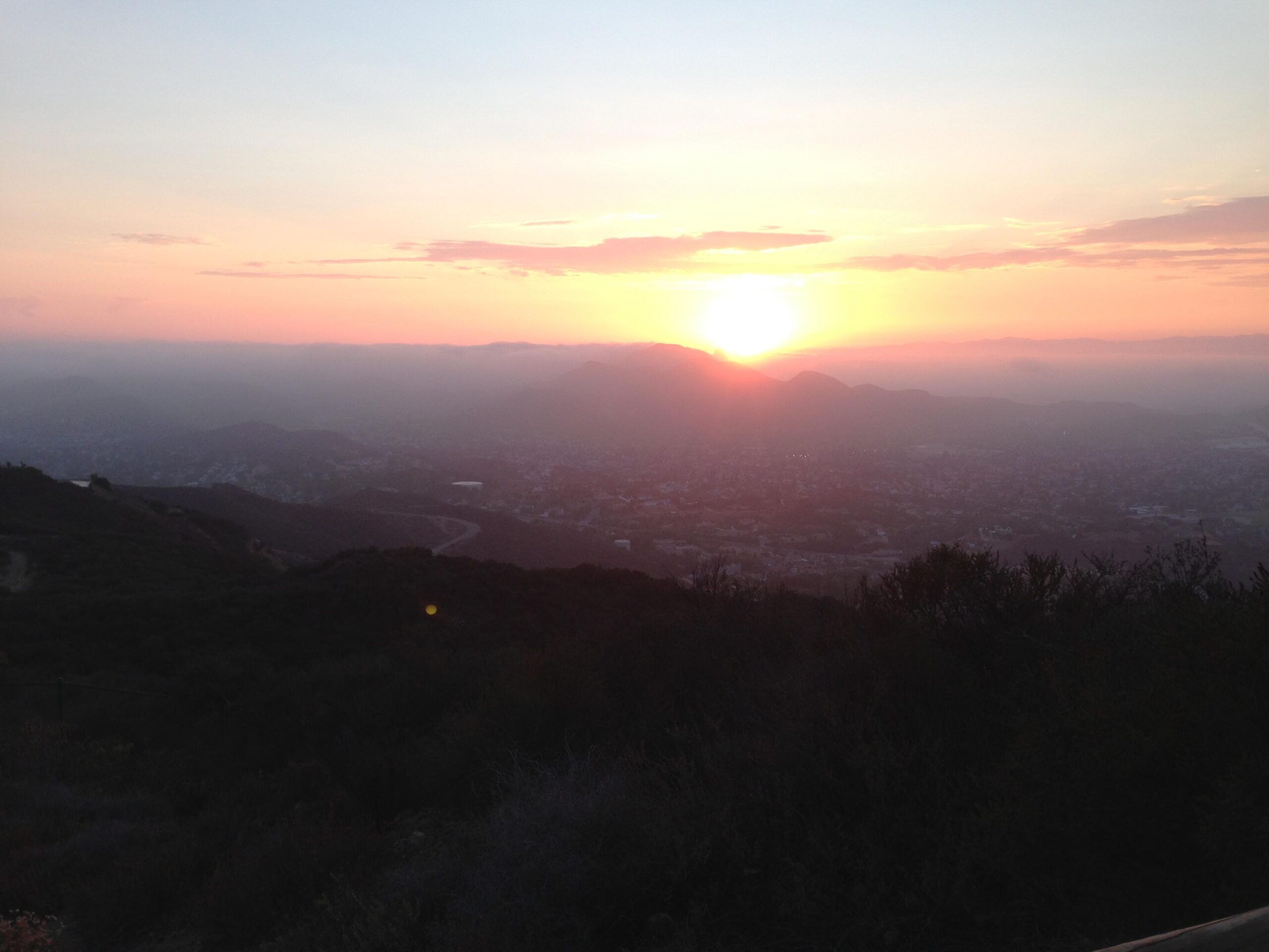 A sunset over a mountainous landscape, with soft pink and orange hues in the sky. The foreground features dark silhouettes of hills and vegetation, while distant city lights twinkle below in the valley. Los Robles Trail (Western Section) mountain bike trail.