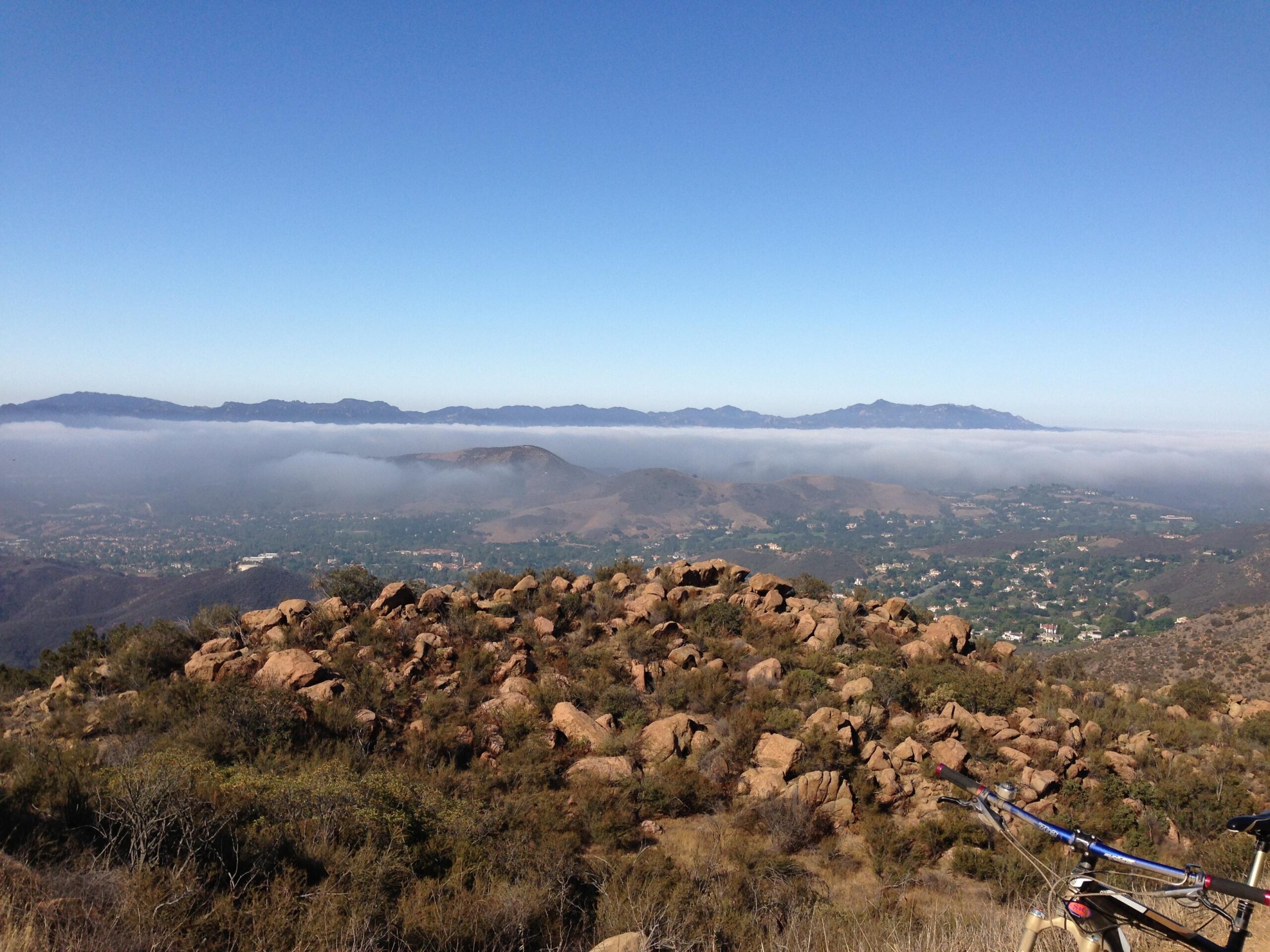 A panoramic view of a mountainous landscape with rocky terrain in the foreground, overlooking a valley covered in fog. The sky is clear and blue, while distant mountains are visible in the background, partially obscured by the mist. A bicycle is positioned at the edge of the rocks, suggesting a scenic outdoor adventure. Cheeseboro / Palo Comado Canyon mountain bike trail.