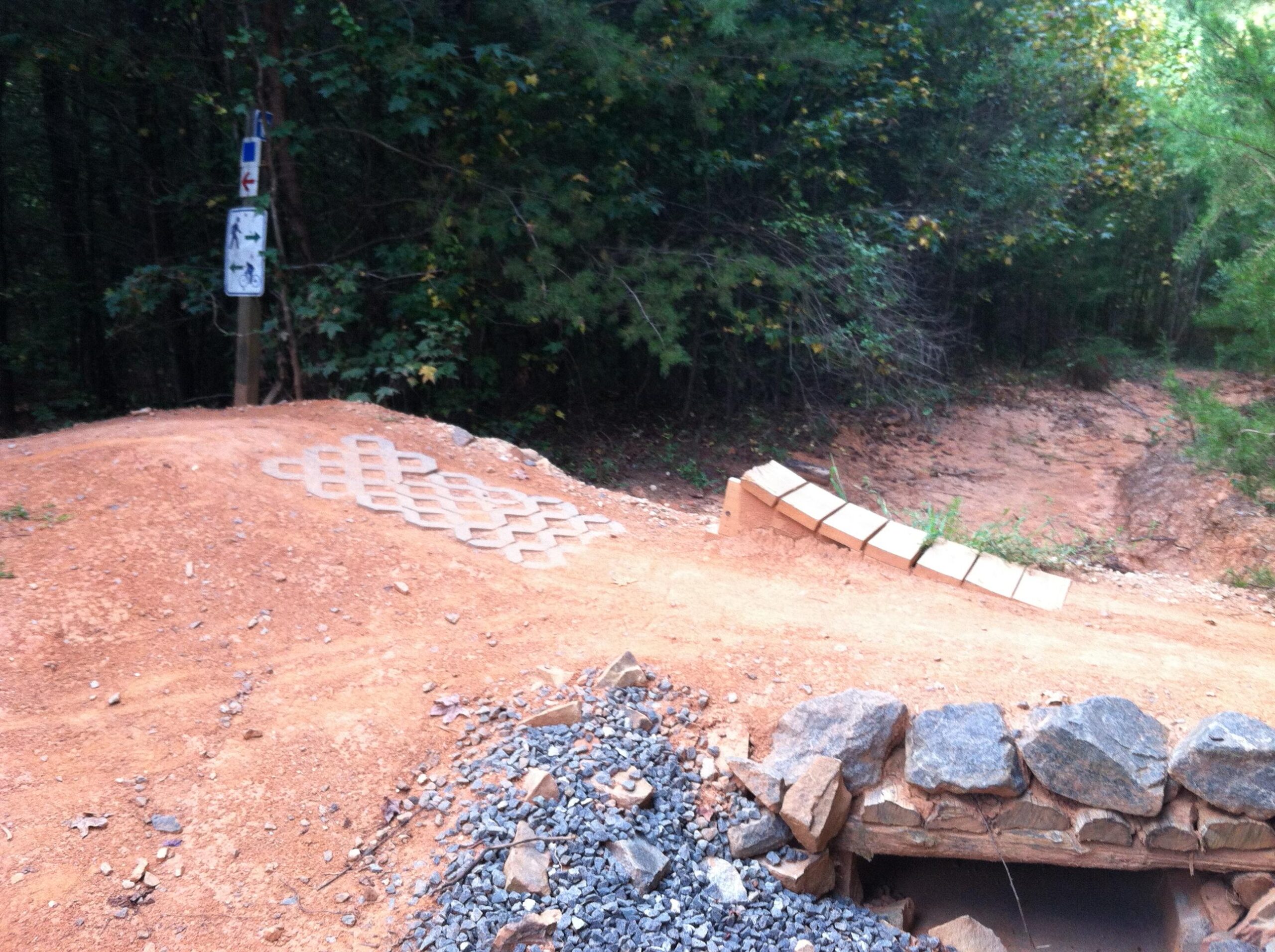 A dirt path leads into a wooded area, featuring a section with patterned paving stones. To the left, a sign indicates directions for trails, with symbols for bikes and pedestrians. A small hill made of dirt and gravel rises on the right, where a row of bricks is arranged at an angle, and a rocky area is visible at the base of the hill. Colonel Francis Beatty Park mountain bike trail.