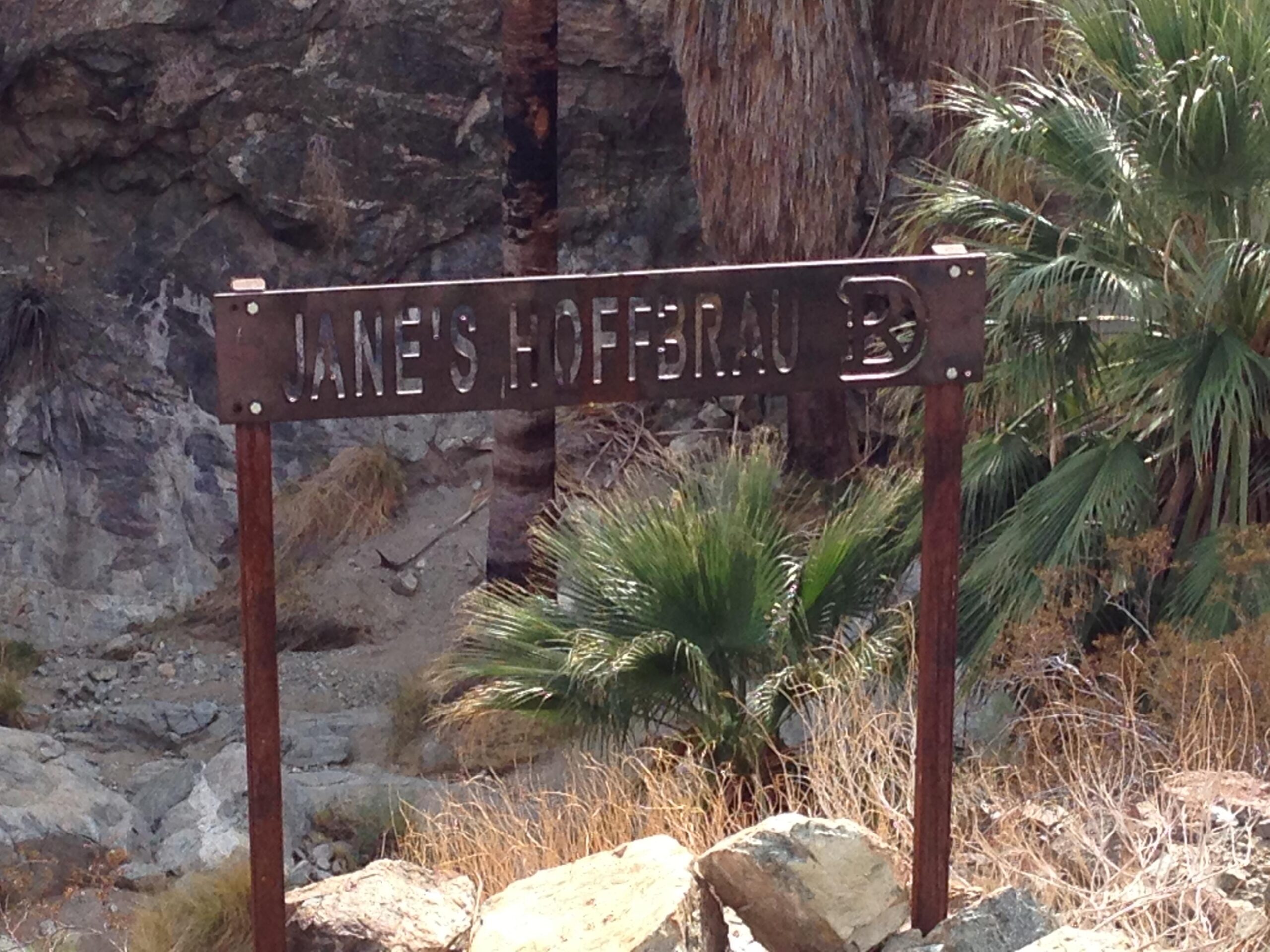 Sign marked "Jane's Hoffbrau" stands amidst rocky terrain and palm vegetation. The Goat Trails mountain bike trail.