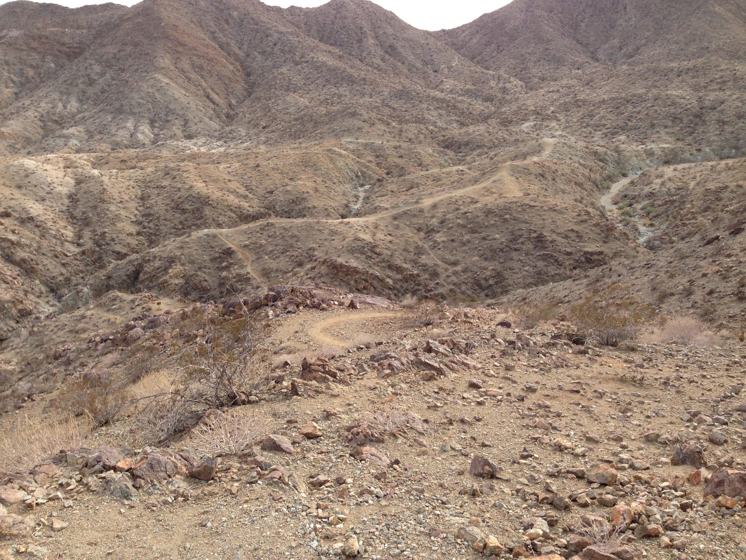 A barren mountainous landscape with rocky terrain and sparse vegetation. Winding paths are visible, leading through the hills and valleys under a cloudy sky. The scene conveys a sense of rugged natural beauty. The Goat Trails mountain bike trail.