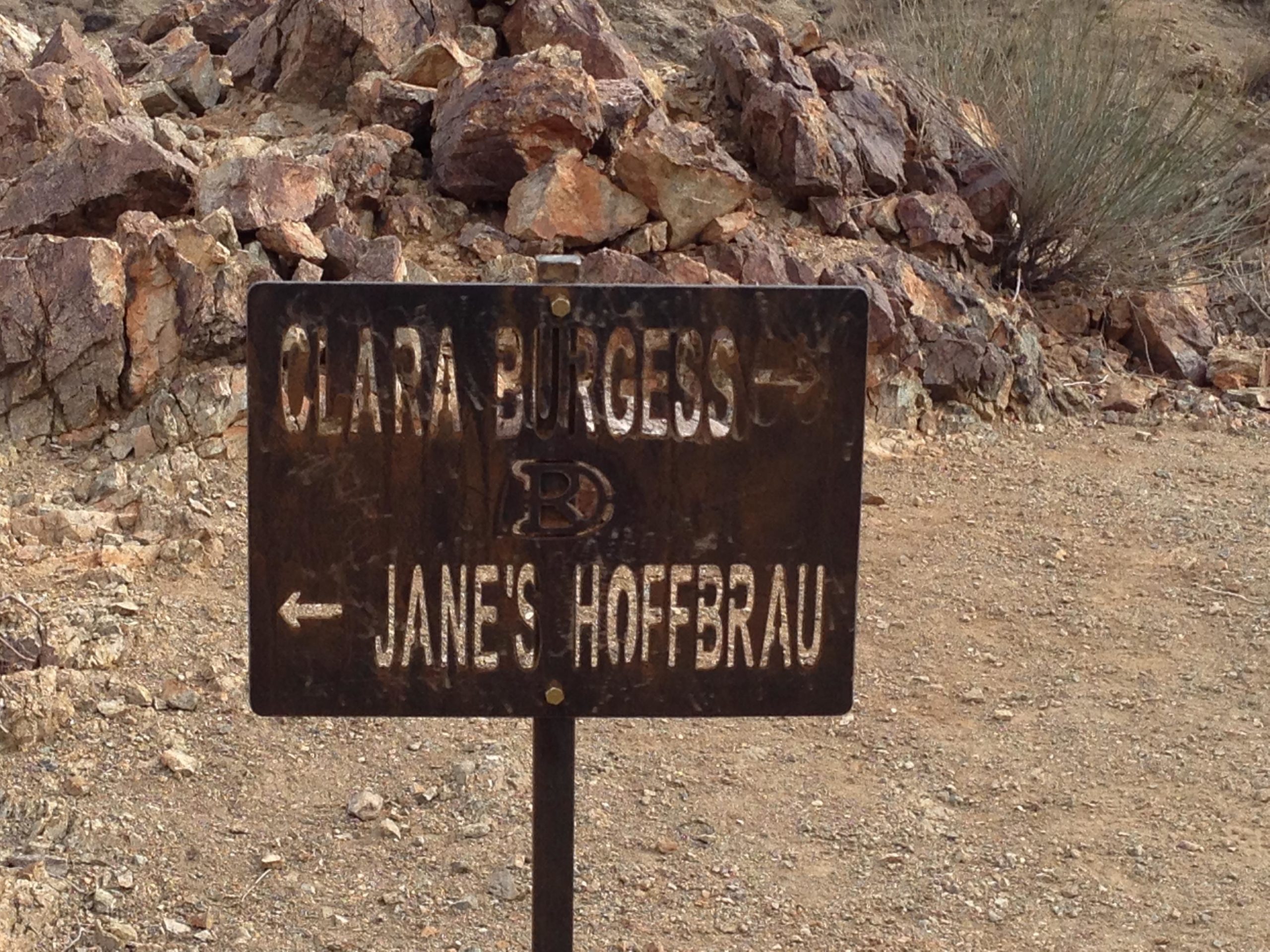 A weathered directional sign indicating two locations: "Clara Burgess" pointing right and "Jane's Hoffbrau" pointing left, set against a rocky terrain. The Goat Trails mountain bike trail.