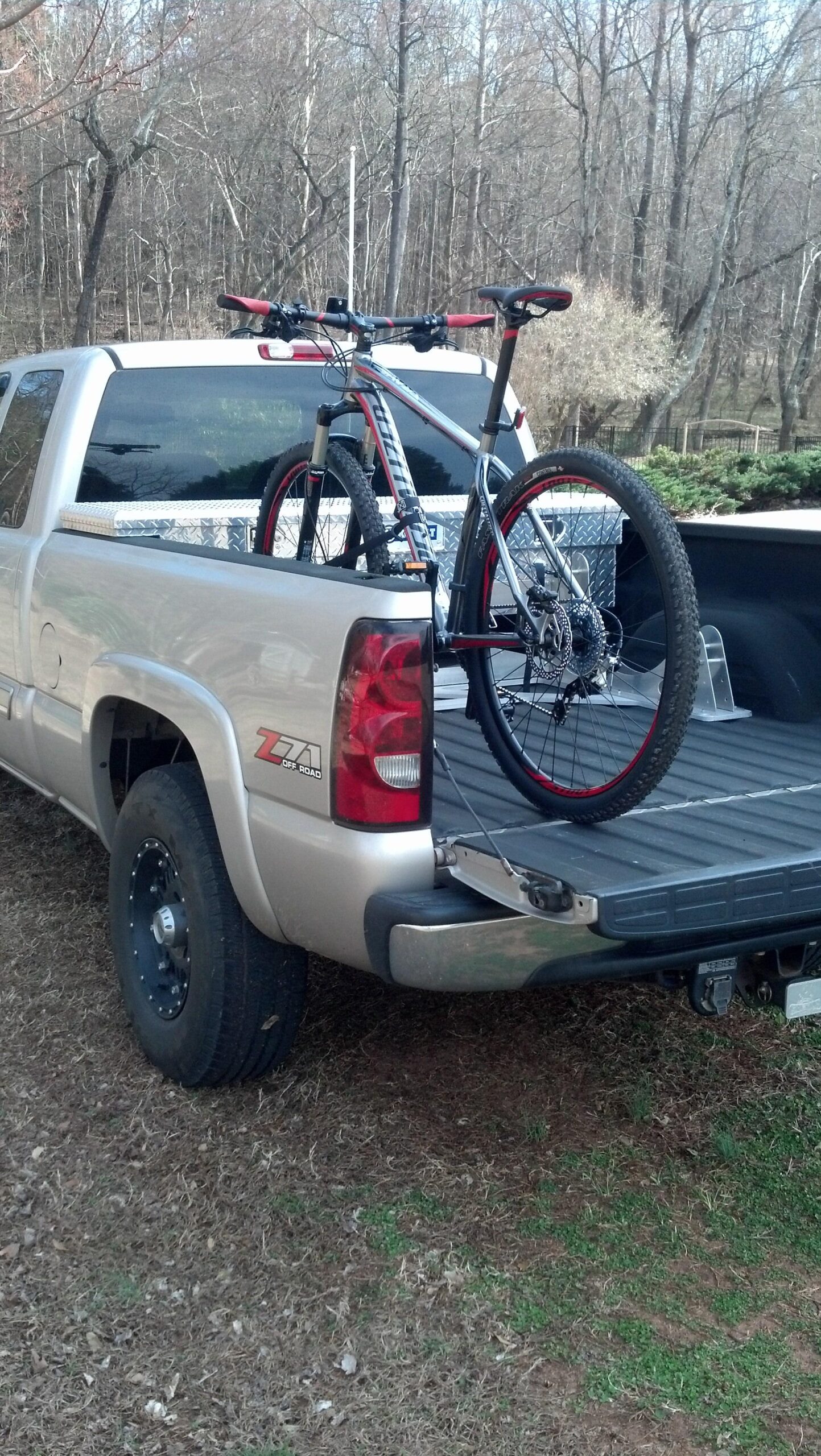 Specialized Crave Expert 29: A mountain bike secured in the bed of a silver pickup truck, with a backdrop of bare trees and grass. The truck has a Z71 off-road emblem and features a dark-colored wheel.