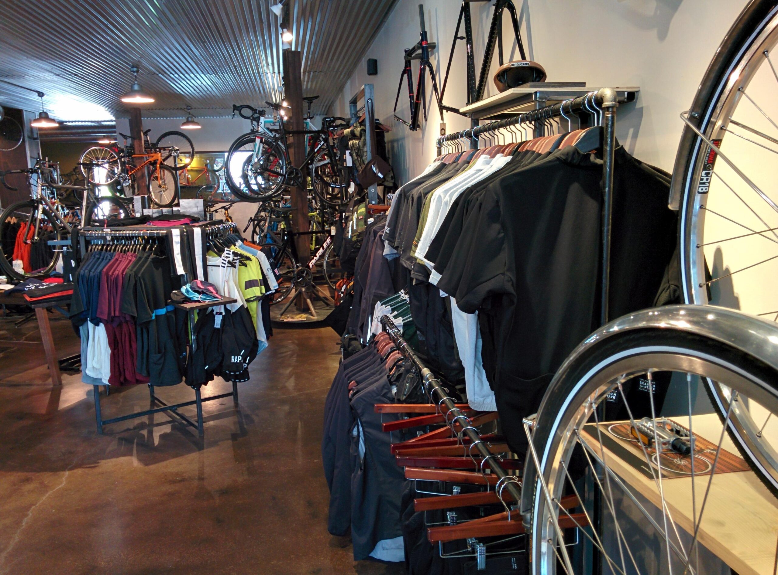 A view of a bicycle shop interior featuring racks of cycling apparel, including jerseys and shorts, displayed on hangers. Various bicycles are mounted on walls and standing on display, emphasizing the shop's focus on cycling gear. The flooring is polished, and the ceiling has a metallic finish, contributing to the shop's modern atmosphere.