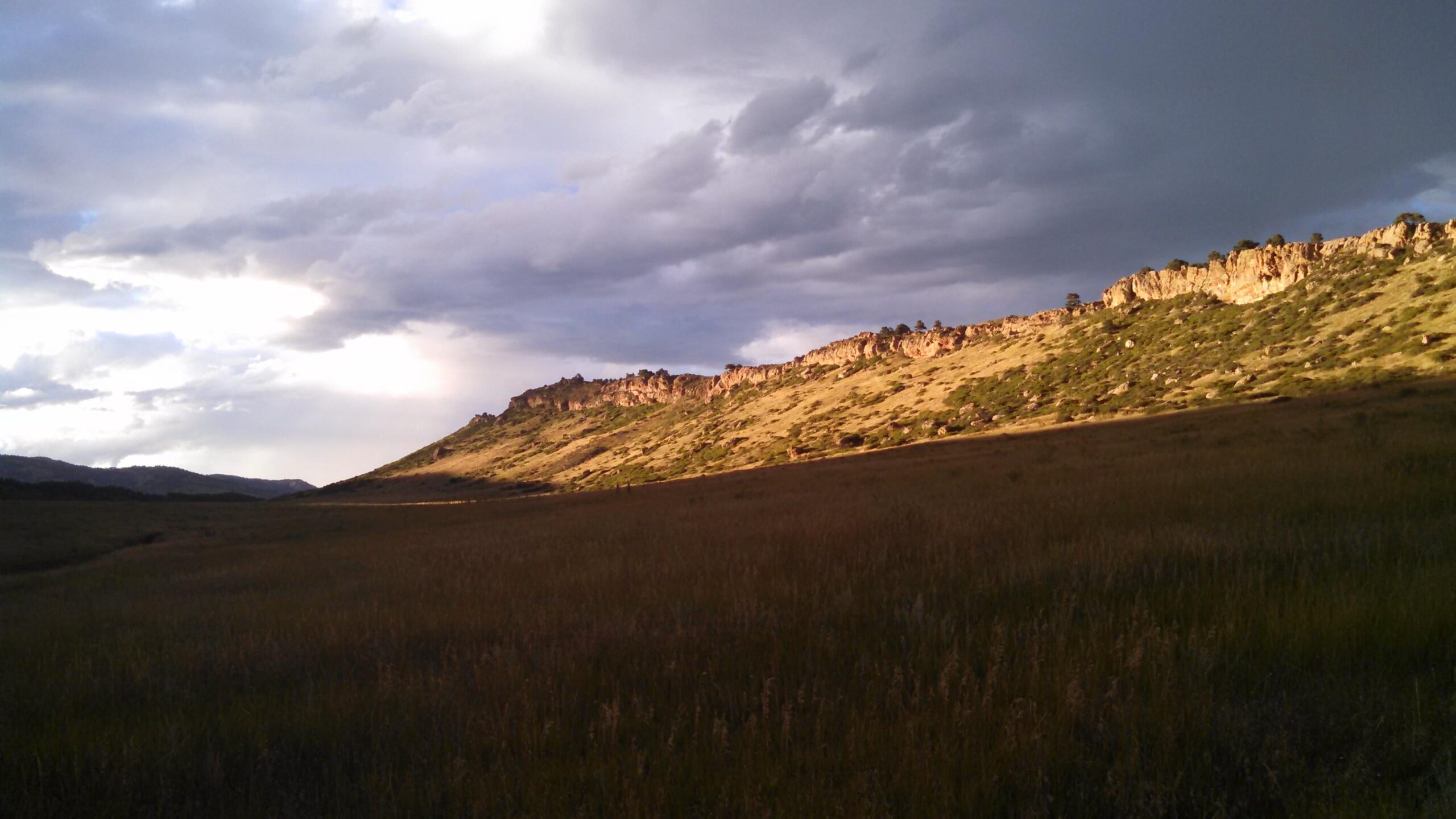 A scenic view of a grassy landscape with rolling hills and rugged rock formations under an overcast sky, featuring patches of sunlight illuminating parts of the terrain. Coyote Ridge mountain bike trail.