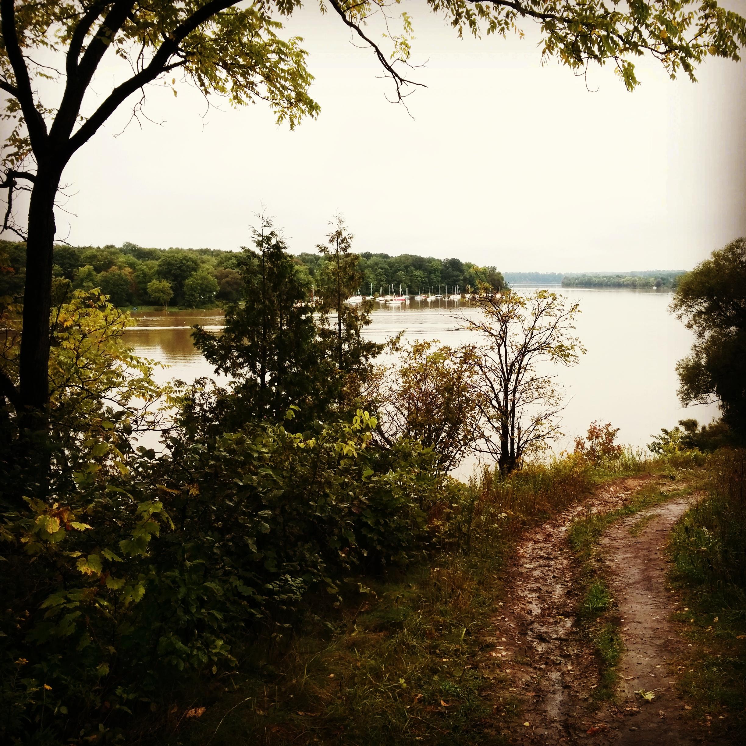 A serene view of a river, surrounded by lush greenery, with a winding dirt path leading towards the water. The sky is overcast, creating a calm and tranquil atmosphere. Boating docks are visible in the distance along the riverbank. Fanshawe Lake mountain bike trail.