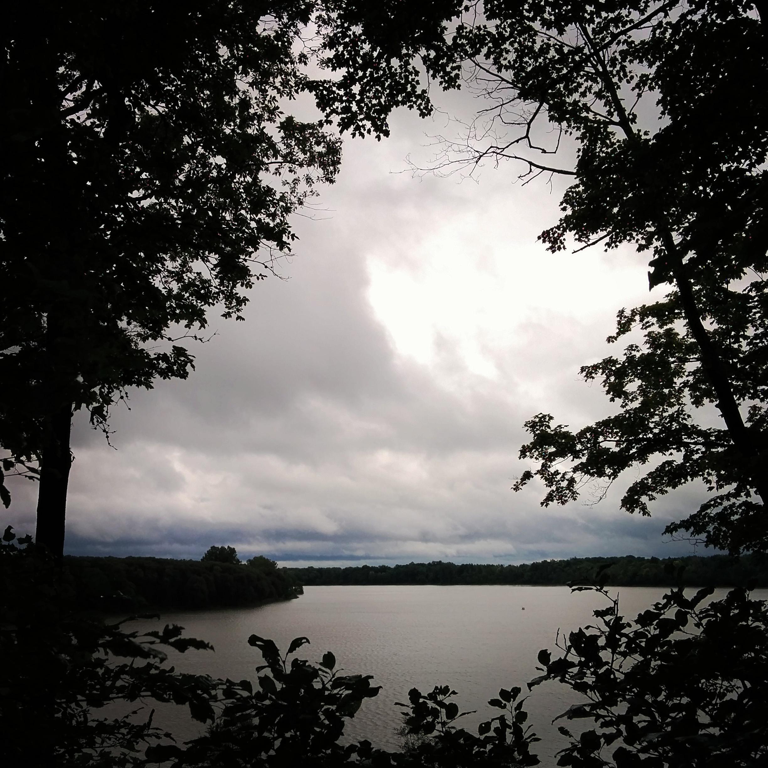 A tranquil view of a lake bordered by trees, with overcast skies. The landscape features calm water reflecting the gray clouds, creating a serene and moody atmosphere. Fanshawe Lake mountain bike trail.