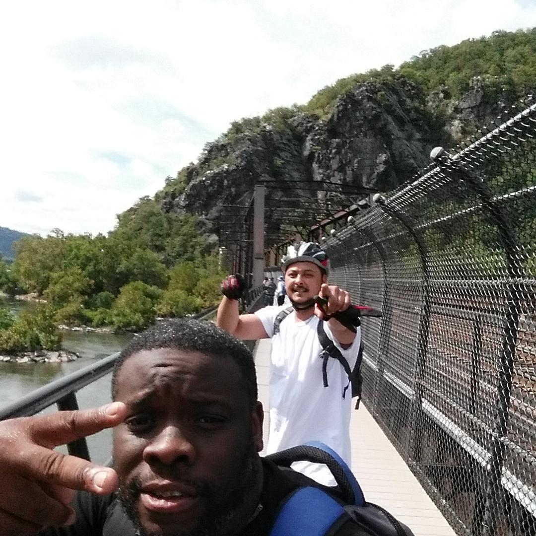 Two people posing for a selfie on a bridge with a scenic view of nature in the background. One person is making a peace sign, while the other is playfully pointing at the camera. A rocky hillside is visible behind them, and the sky is partly cloudy. Both individuals appear to be enjoying a day outdoors. Seneca Ridge Trail mountain bike trail.