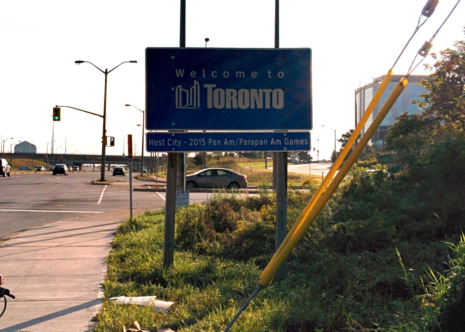 A blue welcome sign for Toronto, with text indicating it was the host city for the 2015 Pan Am and Parapan Am Games. The sign is positioned near a roadway with traffic lights and vehicles, surrounded by greenery and urban infrastructure. Etobicoke Creek mountain bike trail.